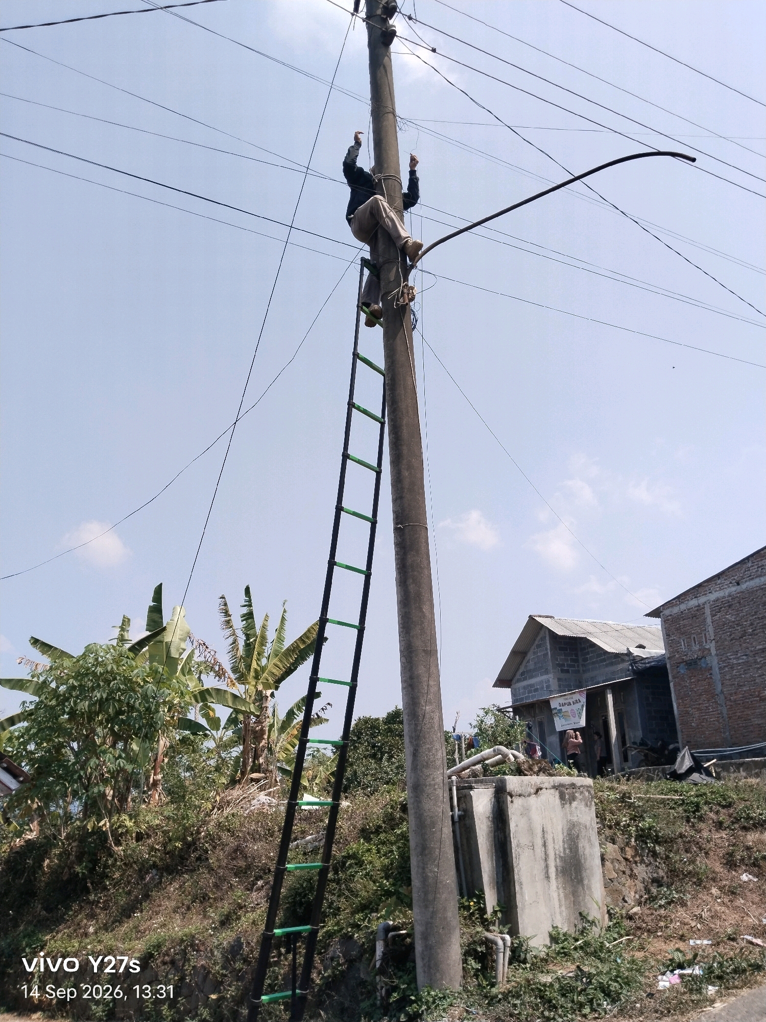 A person is seen climbing a utility pole with a ladder. The person is wearing dark clothing and light-colored pants, with their legs bent and feet on the ladder rungs. Their arms are raised, possibly in the process of working on or adjusting wires. The utility pole is tall and has multiple dark wires extending from it across the clear blue sky. A black, curved rod is also attached to the pole. A green and black ladder is leaning against the pole, extending upwards. In the background, there are trees, including banana trees, and some buildings with corrugated roofs. The scene appears to be in a semi-rural or suburban area. The timestamp "14 Sep 2026, 13:31" and "vivo Y27s" are overlaid on the bottom left corner of the image. The overall impression is one of an outdoor task involving electrical work or maintenance.