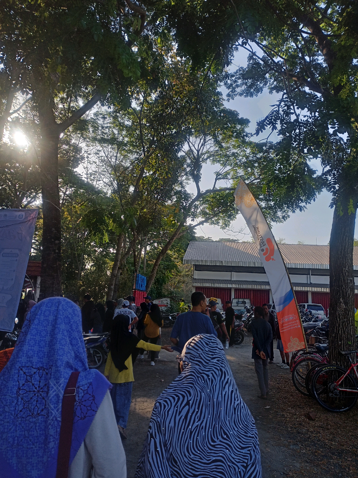 The image depicts a group of people gathered outdoors in what appears to be a public area. In the foreground, three individuals are visible from behind, with two wearing headscarves. One headscarf is blue with a geometric pattern, and the other has a zebra-like stripe pattern. The background shows a building with a red lower section and a corrugated metal roof, suggesting it might be a community center or warehouse. Trees with dense foliage are prominent, casting shadows and allowing sunlight to filter through, creating a dappled lighting effect. Several motorcycles and bicycles are parked in the area, indicating a mode of transport for attendees. A tall, narrow flag with an orange and white design is planted in the ground, featuring text that is partially obscured. People are standing around, some talking, while others appear to be walking. The ground is a mixture of gravel and dirt. The scene suggests an event or gathering taking place in a semi-outdoor setting.