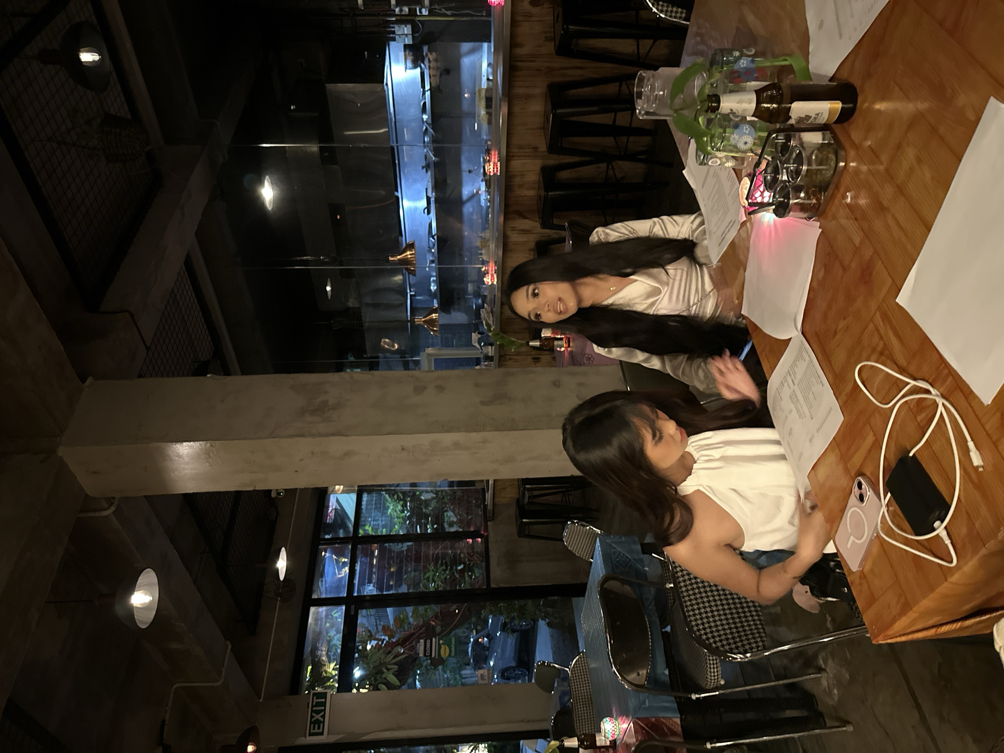 In a dimly lit restaurant interior, two young women are seated at a wooden table. The woman on the left wears a white halter-neck top and is looking down at papers in front of her. The woman on the right wears a satin-like light-colored blouse and is looking to her left. On the table are several papers, a phone, a portable charger with cables, a set of spice jars, a glass pitcher of water, a bottle of beer, and a small decorative candle. In the background, a kitchen counter with stainless steel appliances is visible. The restaurant appears to be located in Davao City, Philippines, as indicated by a "GrabFood" sticker on the window in the background. The overall ambiance is casual and social.
