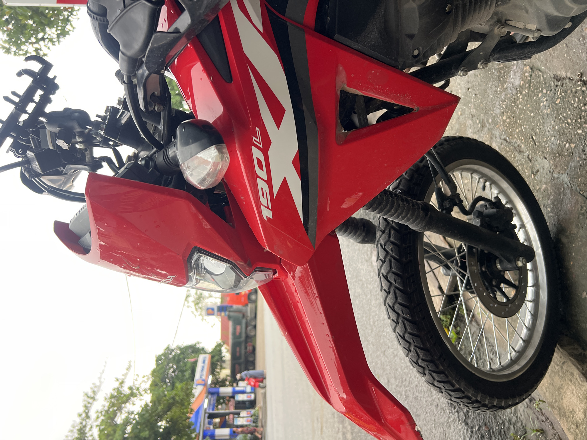 The image shows a red Honda 190L motorcycle parked outdoors on a wet asphalt surface. The motorcycle is facing right, with its front wheel and part of the fairing in focus. In the background, a gas station is visible, with pumps and a canopy. There are also a few people walking and a large truck is parked behind the gas pumps. The scene appears to be in Nông Tiến Ward, Vietnam. The sky is overcast, suggesting it might be raining or has recently rained.