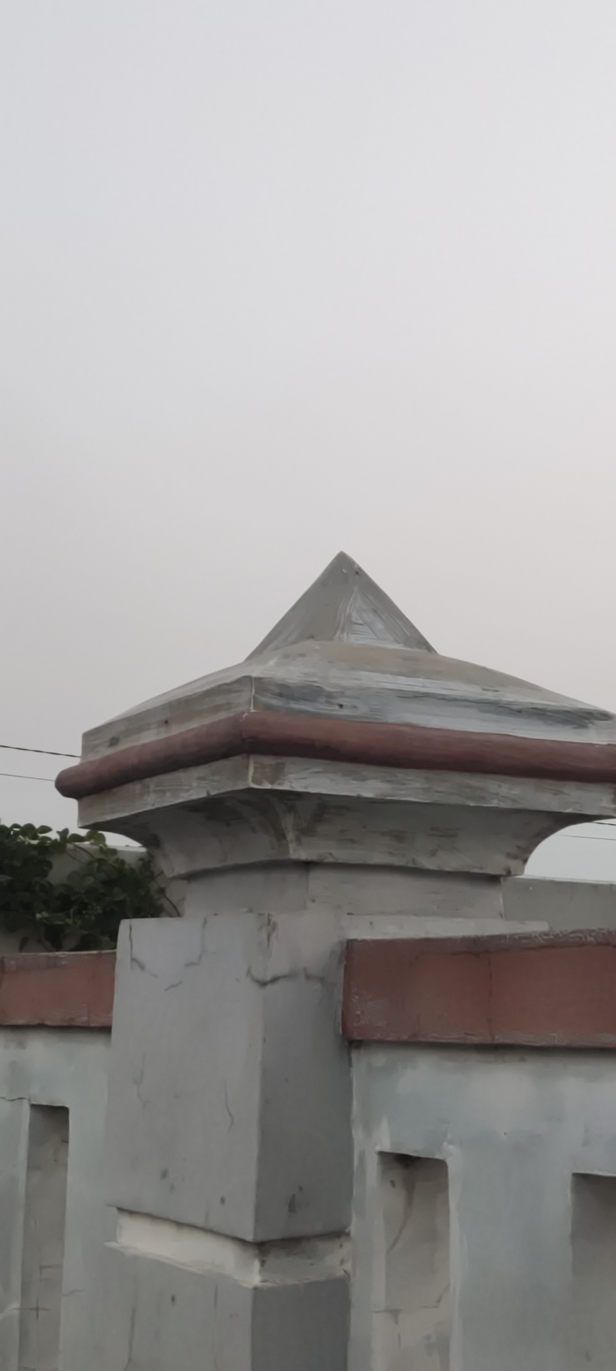 The image shows a close-up of a concrete fence post topped with a pyramid-shaped cap. The cap is painted white with some weathering and a reddish-brown band around its base. The fence itself is made of concrete with decorative rectangular cutouts and a reddish-brown trim. In the background, there is a hazy sky and some green foliage is visible to the left. The scene appears to be outdoors. No people are present in the image.