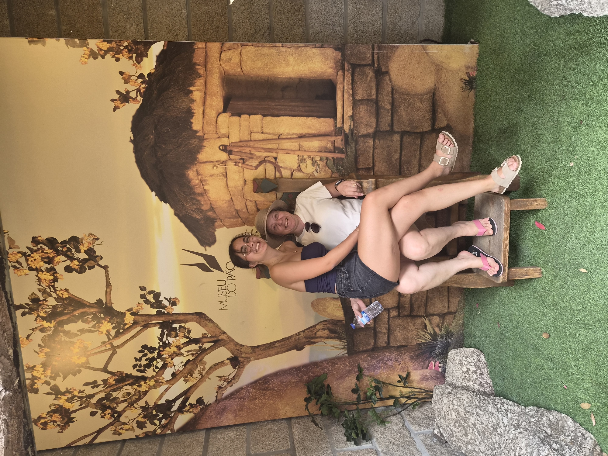 Two individuals are posing in front of a large, printed backdrop that depicts a rustic scene with a thatched-roof dwelling, a tree with yellow flowers, and mountainous terrain. The backdrop also features text that reads "MUSEU DO PAO". The setting is in front of stone walls.
The person in the foreground is a young woman with dark hair, wearing glasses, a dark blue strapless top, and denim shorts. She is sitting on the lap of the person behind her, holding a plastic bottle of water in her right hand. She is smiling and looking at the camera.
The person behind her is an older woman with blonde hair, wearing a white t-shirt and a straw hat. She is also smiling and looking at the camera. She is wearing sandals.
The ground in front of them is covered with artificial green grass, and there are a few fallen leaves scattered on it. There are also some large rocks to the left of the scene. The overall impression is of a playful photo opportunity at the Museum of Bread (Museu do Pão) in Seia, Portugal.