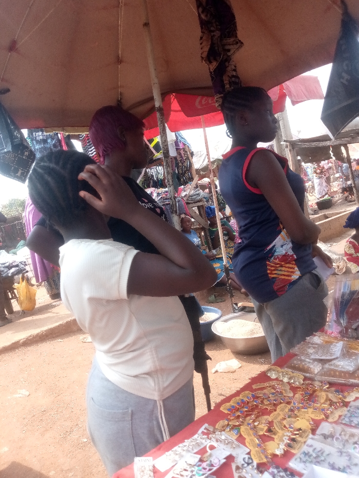 The scene depicts an outdoor market in Kpak, Nigeria. Three females are in the foreground and midground. On the left, a female with dark braided hair, wearing a light top and light grey pants, stands with her right hand touching her head. Center-right, a female with short purple hair, wearing a dark t-shirt, stands in profile facing right. To her right, another female with dark braided hair, wearing a navy blue sleeveless top with a colorful design and dark grey pants, also stands in profile facing right, with her hands clasped in front.
The immediate foreground features a table covered with a red cloth, displaying various small items for sale, including necklaces, earrings, rings, and keychains, many individually packaged on cards or in clear plastic. Above the subjects, a light brown canopy provides shade, supported by metal poles. Clothing and bags, including one with "Emirates" branding, hang from these poles. In the background, a red Coca-Cola umbrella is visible.
On the reddish-brown dirt ground, two silver metal bowls contain light-colored granular material, and a blue plastic bowl is nearby. Other blurred figures and market structures are visible in the background, indicating a busy environment. The lighting suggests a bright day.