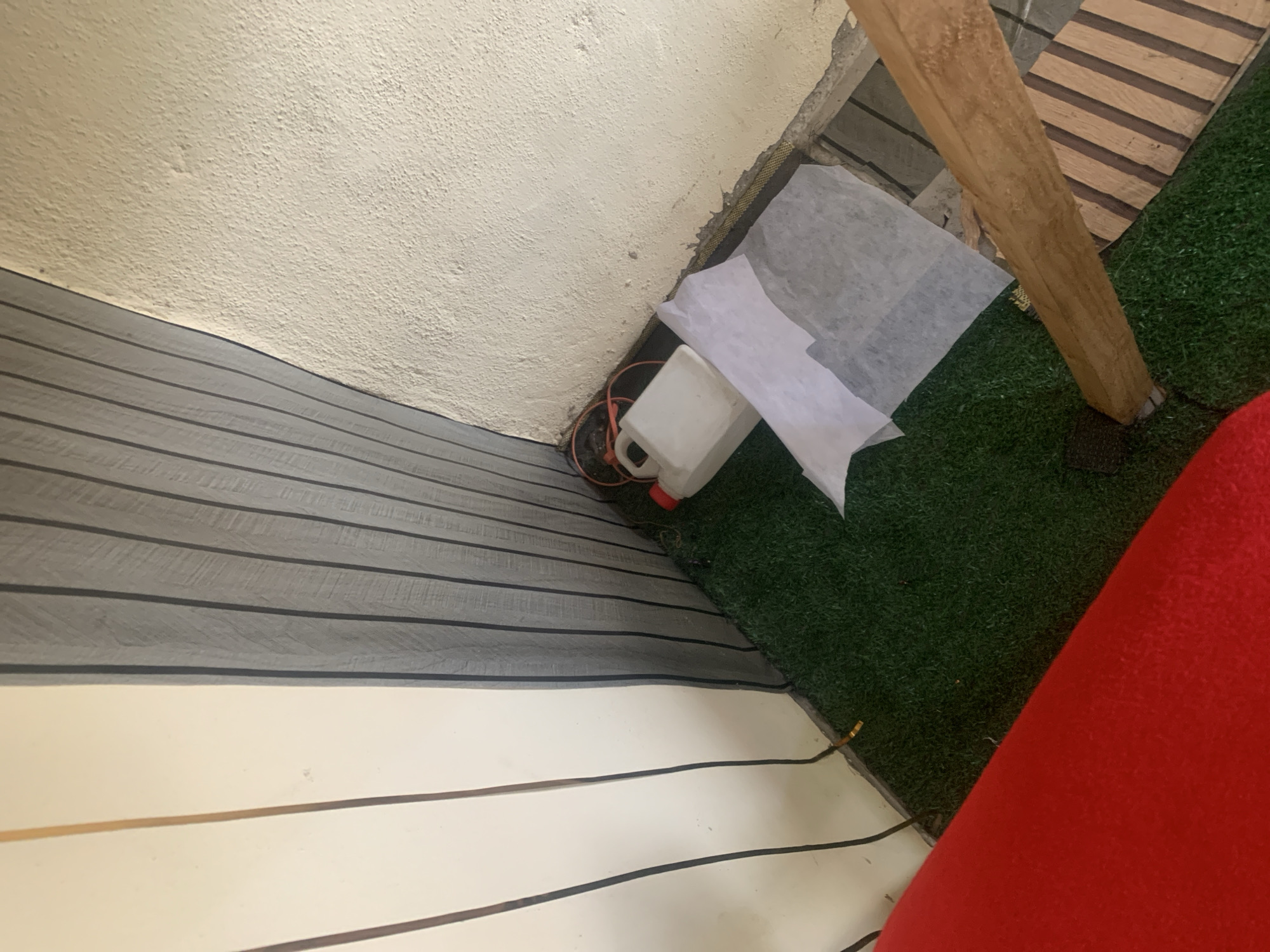 The image shows a corner of an outdoor area with artificial turf flooring. A white plastic container with an orange lid and orange wires is situated on the turf. A white sheet of paper or fabric lies partially over the container. To the right, a wooden beam is propped diagonally across the scene, resting on the turf and extending towards a textured wall. The walls are a mix of vertical grey paneling and a rough, cream-colored stucco. The overall setting suggests an unfinished or storage area. There are no people visible in the image.