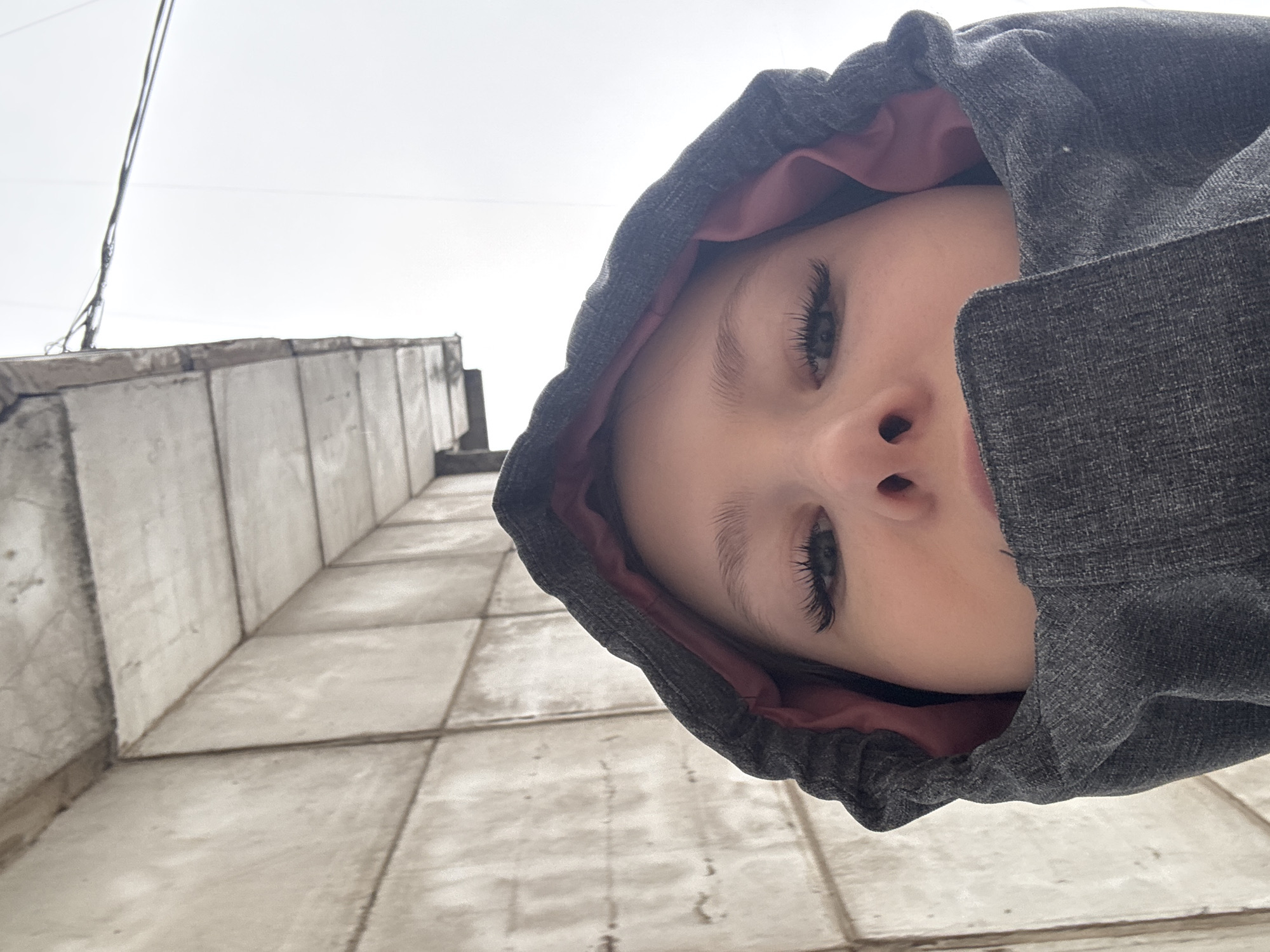 A young person wearing a grey hooded jacket with a pink lining is positioned in the foreground, their face tilted towards the camera, filling the upper-right portion of the frame. The background reveals a concrete building exterior with a tiled surface. A utility wire is visible against the overcast sky in the upper-left corner. The setting is likely in an urban area, potentially Chaikovsky, Russia.