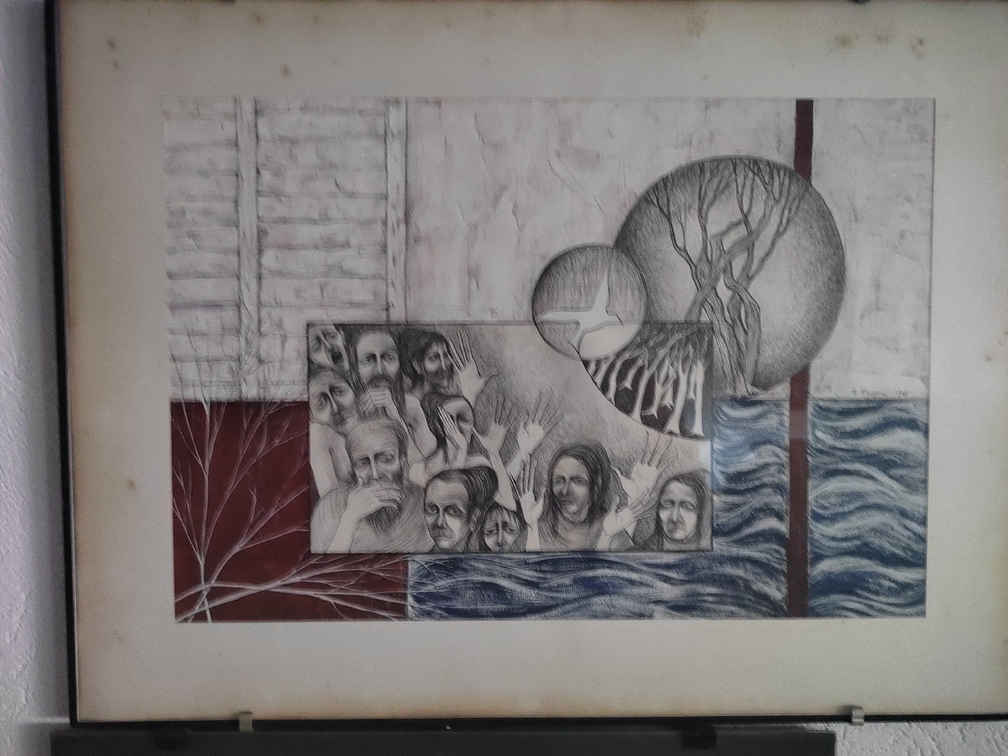 This is a photograph of a framed charcoal and pastel drawing. The drawing depicts a surreal scene with several distinct elements. On the left, there is a textured background resembling brickwork or wood paneling, with a stylized, bare tree branch motif in white and red against a dark red rectangular field. To the right of this, a cluster of human faces is rendered in charcoal, some with expressive emotions like distress or shock. Several figures have their hands raised. Above this group of faces, two overlapping circles are drawn. The larger circle contains a detailed drawing of a gnarled tree, its branches reaching outward. The smaller, overlapping circle seems to contain a stylized bird or winged figure. The right side of the drawing features a wavy, abstract representation of water in shades of blue and white, bordered by a vertical dark red line. The overall composition is complex and evocative, with a somber and dreamlike quality. The artwork is housed in a light-colored mat and a dark frame. The photograph is taken from a slightly elevated angle, capturing the artwork on a wall, with part of a dark frame or easel visible at the bottom. There are some dust specks or imperfections on the matting. There is no indication of a specific location in Porto Recanati, Italy within the image itself.