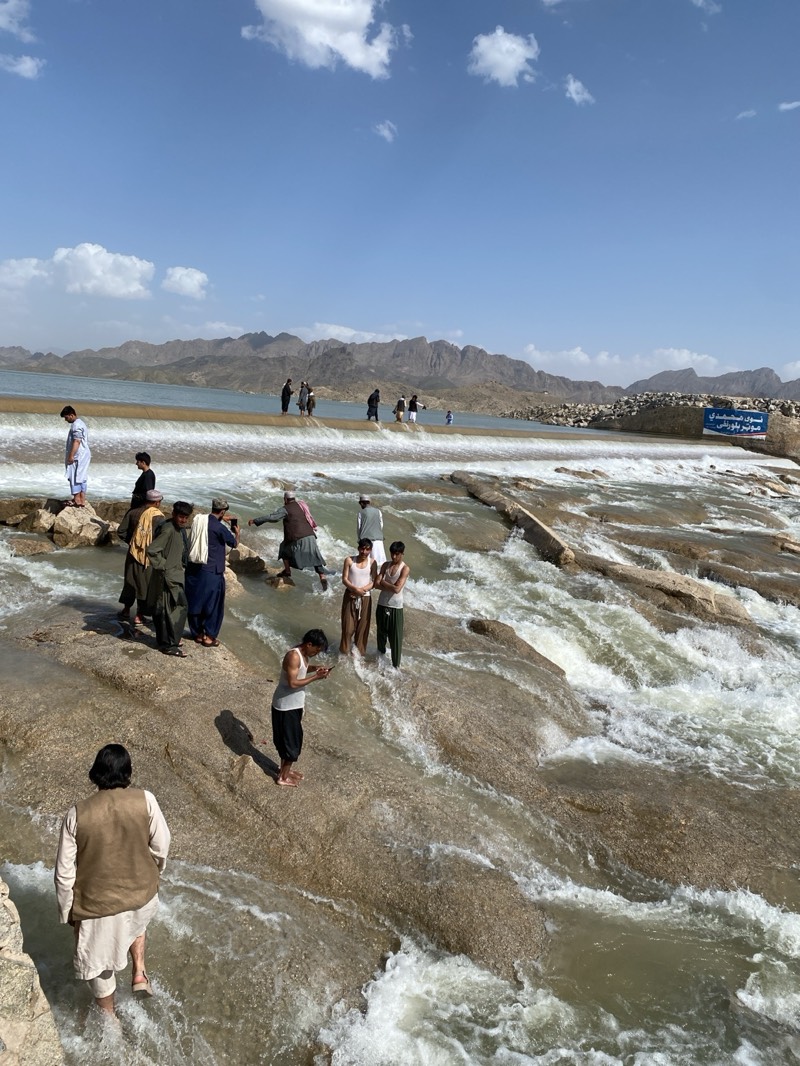 The image shows a group of people at a dam or weir, with mountains in the background under a bright blue sky with scattered clouds. The location is identified as Kim, Afghanistan. Several men are standing in and around the water flowing over the structure. Some are fully clothed, while others are shirtless or partially dressed. One man is looking at a phone. The water is rushing over the stepped stone surface of the dam, creating white water. A sign is visible on the right side of the structure, with text in a non-English script.