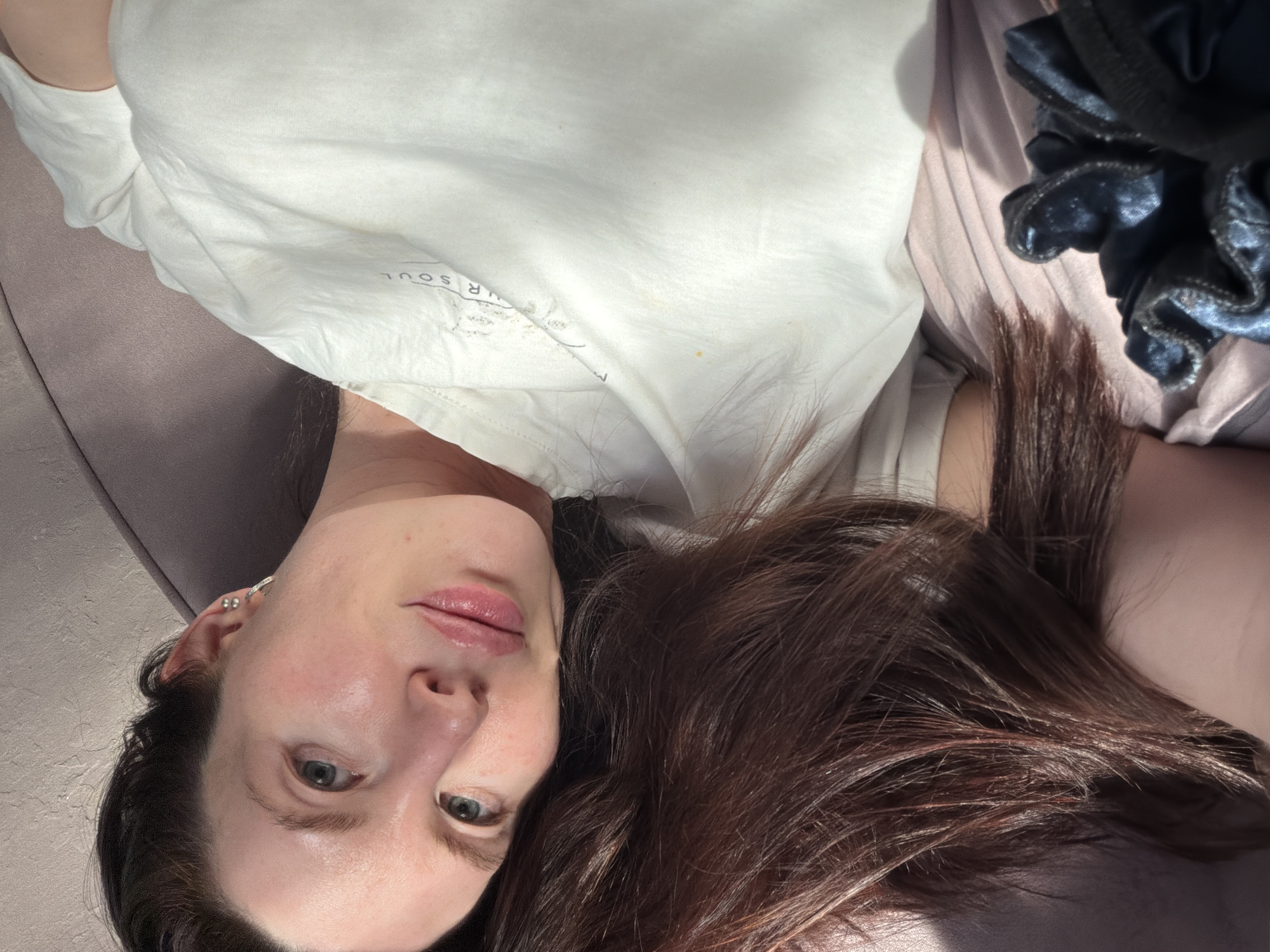 A woman with fair skin and dark brown hair lies on a light purple surface, presumably a couch or bed. She is wearing a white t-shirt. A dark blue scrunchie is visible near her right shoulder. The background is a blurry mix of light and shadow. The image appears to be taken indoors in natural light. The location in Perm, Russia is not evident.