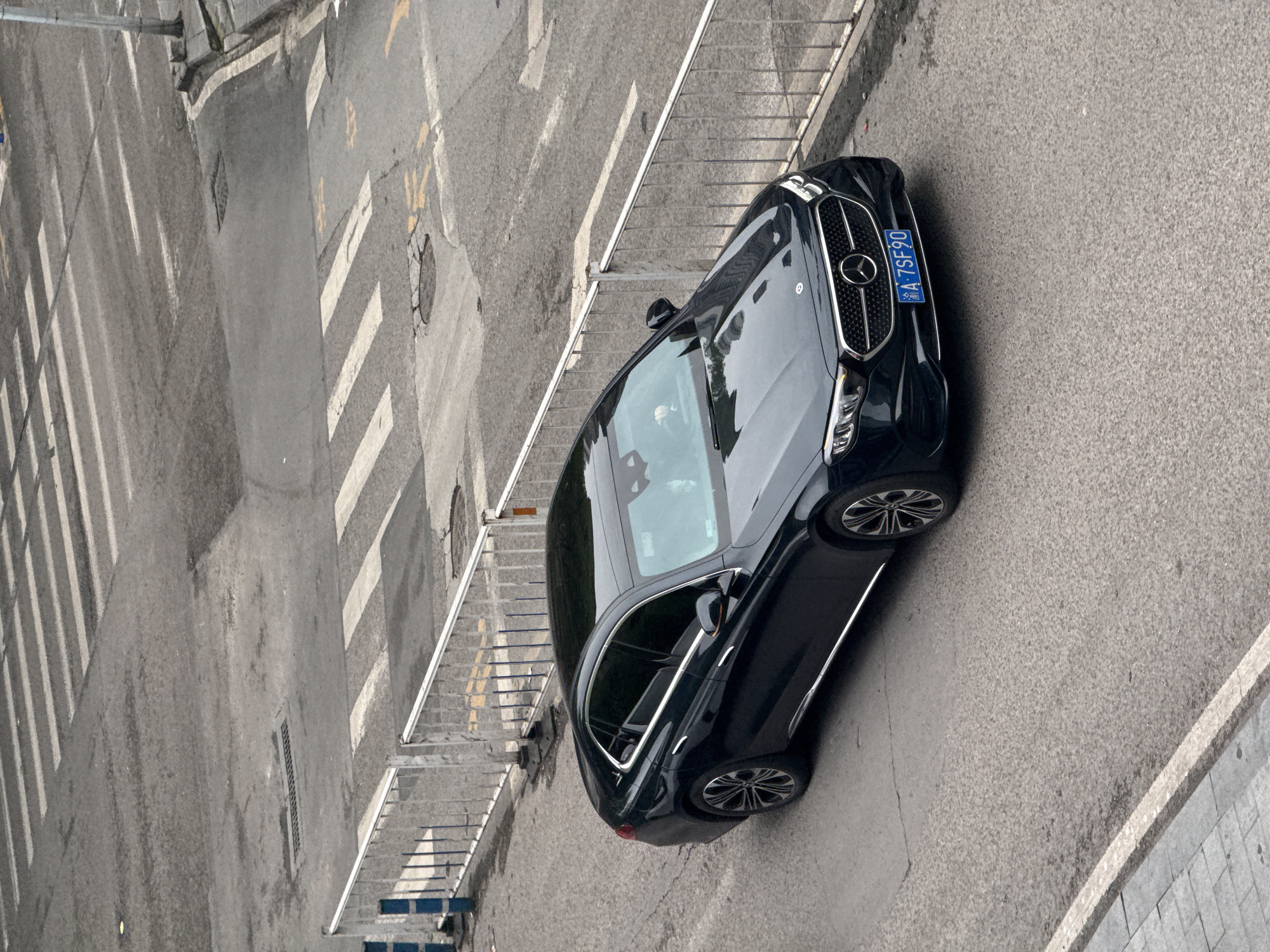 The image shows a black Mercedes-Benz sedan parked on a grey asphalt road in Yuzhong District, China. A crosswalk is visible adjacent to the car, as is a metal railing and a concrete sidewalk. The vehicle's license plate reads "A 7SF90." The image appears to be taken from an elevated position, providing a top-down view of the car and the surrounding urban environment. No people are visible.