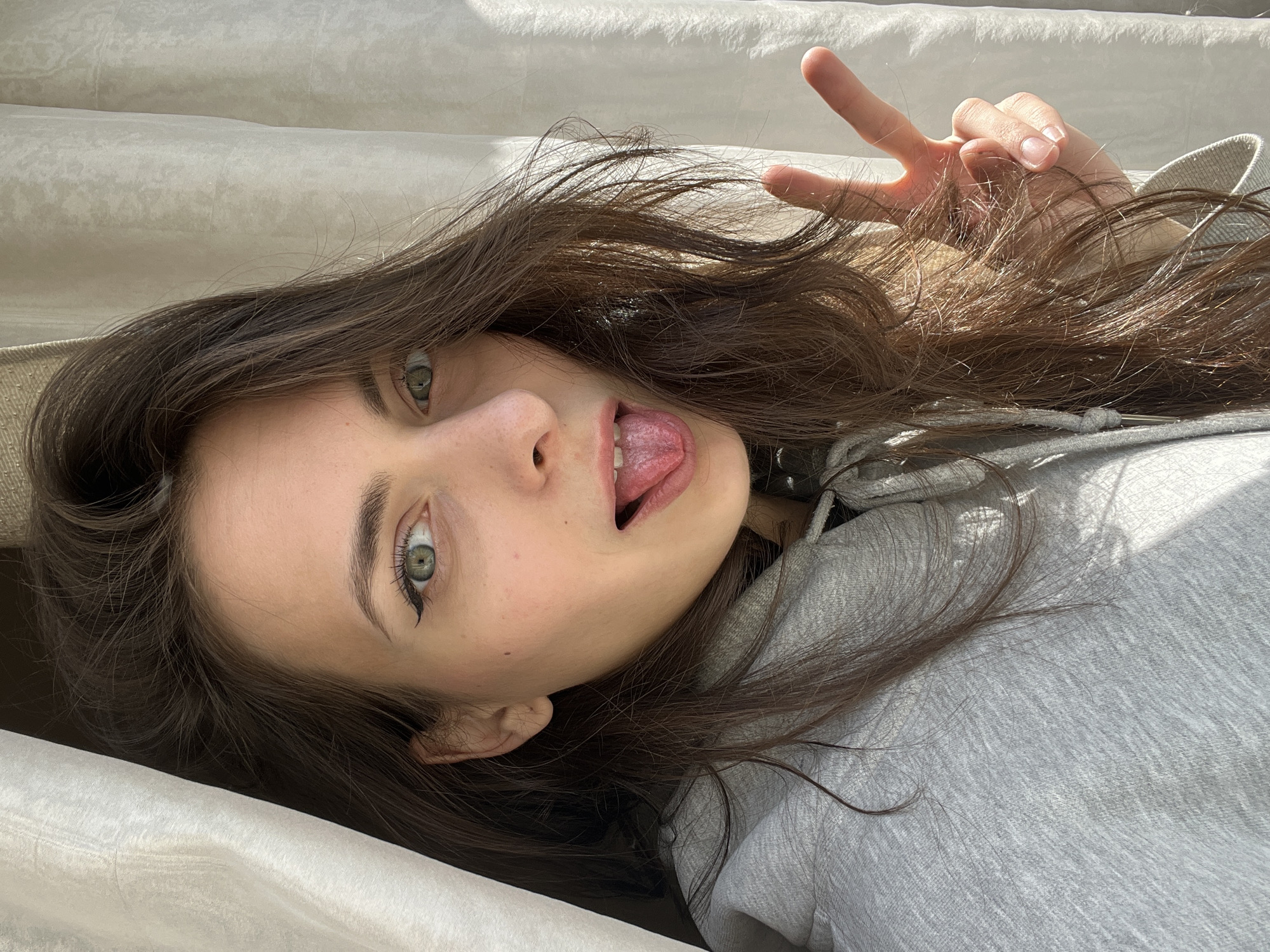 A young woman is lying on a surface of white fabric, possibly a bed or sofa. She is wearing a grey hoodie. Her dark brown hair is spread around her head. She is sticking her tongue out and making a peace sign with her right hand. Her eyes are green. The photo is taken from a close-up, tilted perspective. The location in the city of Krasnoyarsk, Russia, is not discernible from the image.