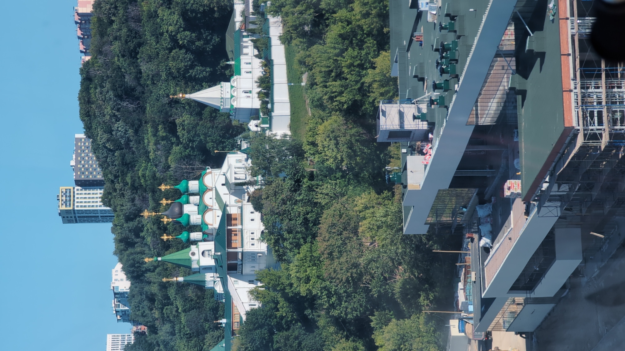 The image depicts an exterior view of a complex featuring orthodox churches with green domes and golden crosses, nestled amongst lush trees. To the left of the churches, several modern high-rise buildings with a predominantly white and grey facade are visible, suggesting an urban or semi-urban setting. The sky is a clear, bright blue, indicating fair weather and likely daytime. The scene is captured from a slightly elevated perspective, looking down upon the churches and into the surrounding greenery. On the right side of the image, a large, modern building with multiple levels and a dark green roof is partially visible, with scaffolding and construction materials hinting at ongoing work or development. The overall impression is one of architectural contrast between traditional religious structures and contemporary urban development, set against a backdrop of nature. No people or text are clearly discernible.