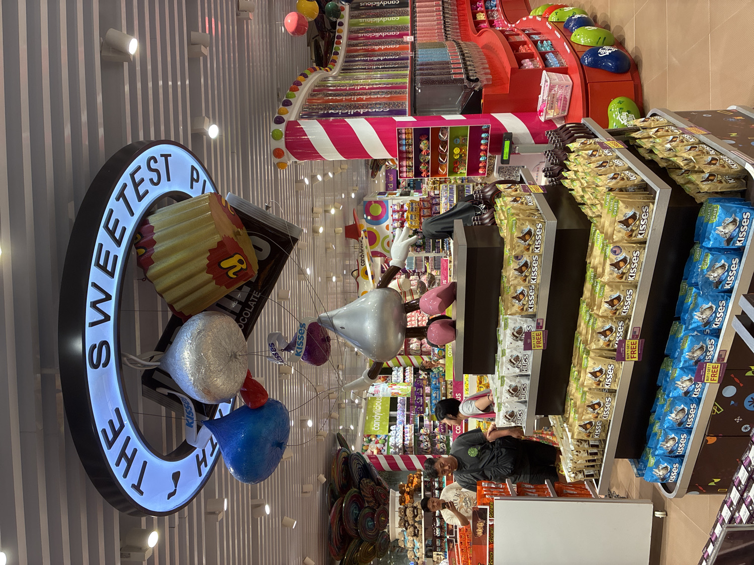 This media file presents a vibrant, well-lit interior of a large candy store, located in Kuta, Indonesia. The scene is dominated by numerous candy displays and oversized confectionery models.
On the left, a prominent circular sign glows with white light, bearing the partially visible text "THE SWEETEST PLA" (likely "THE SWEETEST PLACE"). This sign is integrated with large, three-dimensional models of candies, including a Reese's Peanut Butter Cup with "Reese's" text, a Hershey's chocolate bar (partially showing "OCOLATE"), and several metallic Hershey's Kisses models in silver, red, blue, and purple, appearing to float.
The right and central portions of the image are filled with shelves stocked with various candies. Bags of Hershey's Kisses and Hershey's Hugs are extensively displayed, many featuring yellow "BUY 1 GET 1 FREE" promotional stickers. Some Kisses bags also show "US" text, indicating their origin. Further into the store, tiered displays hold colorful bulk candies, possibly Jelly Beans, and branding for "candylicious" is visible on several shelves.
Several people are present in the store, engaging in typical retail activities. A person in a dark shirt, possibly staff, is seen tending to the Kisses display, while other shoppers or employees are partially visible in the background. The store features a modern design with striped walls and a ceiling equipped with numerous recessed spotlights, suggesting a bright, welcoming commercial environment during daytime hours.