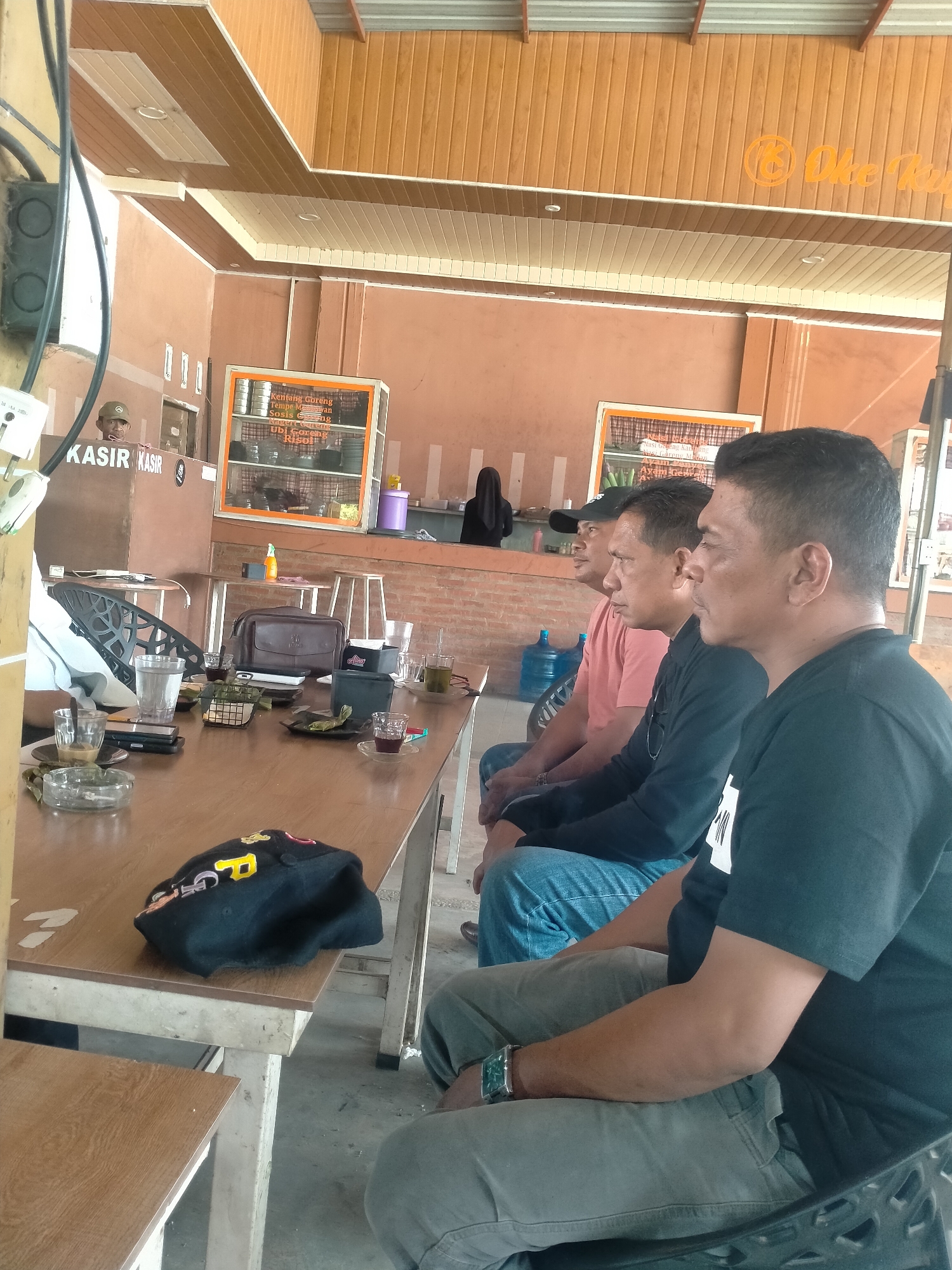 Three men are sitting in chairs at a wooden table inside a building. The men are facing towards the left side of the image. The closest man is wearing a dark t-shirt, gray pants, and a watch. The second man is wearing a dark long-sleeved shirt and blue jeans. The third man is wearing a pink t-shirt and blue jeans, and a black baseball cap. On the table are several glasses, an ashtray, cell phones, a wallet, and a black baseball cap. In the background is a cash register with a man standing behind it. There is also a woman in a black hijab in the background, and a water jug. There are brown wooden walls and ceiling. One sign visible states "Oke Rupat". Another says "Kasir". The menu is visible above the counter. The location is Matang, Indonesia.