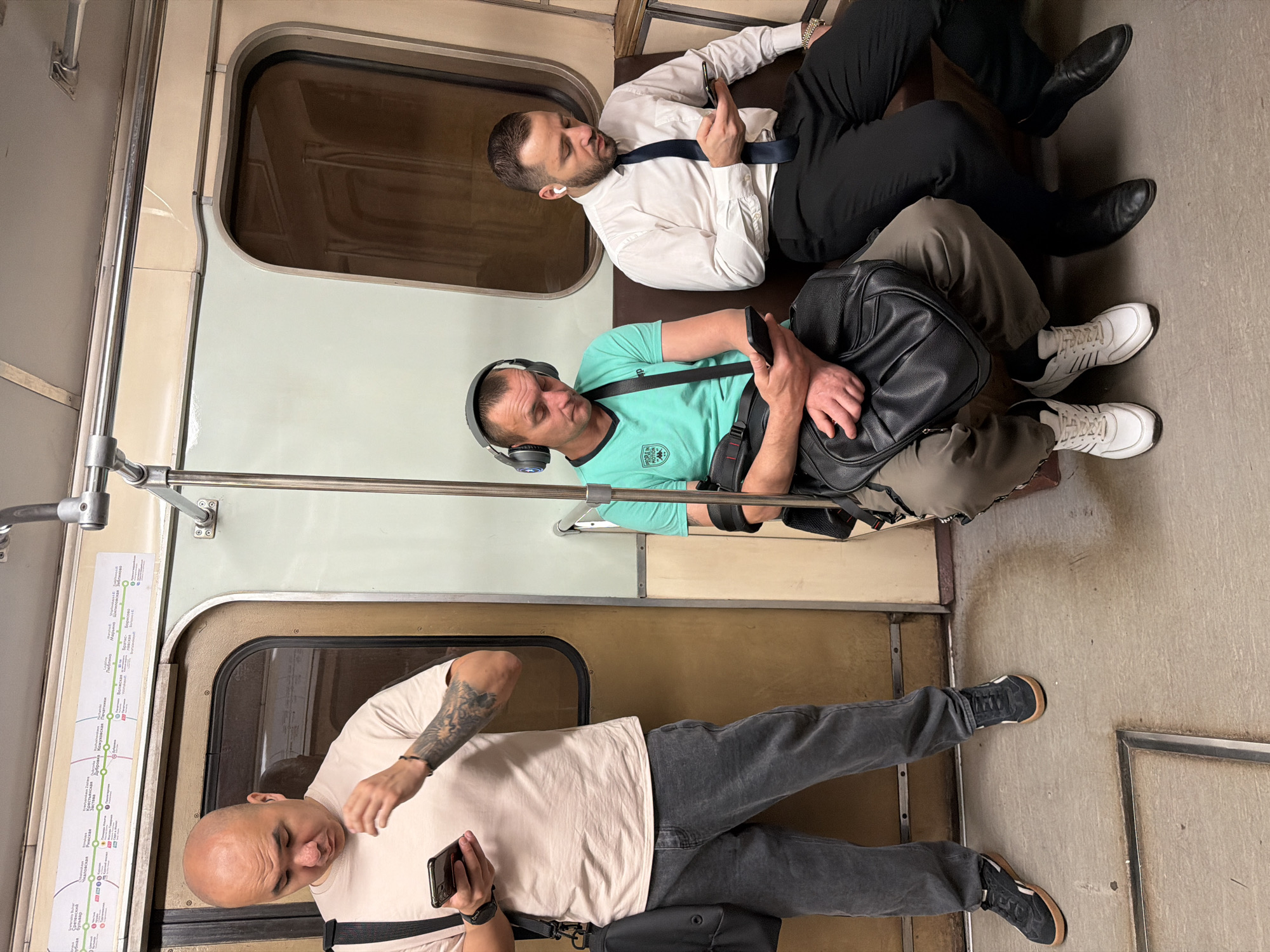 Three men are depicted inside a Moscow Metro train car. On the left, a bald man stands while looking at his phone. He wears a light-colored t-shirt, dark gray jeans, and black sneakers. A black bag hangs over his left shoulder. In the middle, a man with short brown hair and headphones sits, also looking at his phone. He is wearing a teal t-shirt and olive-colored pants, and has a black bag on his lap. To the right, a man with short dark hair and a beard sits, wearing a white collared shirt, dark tie, and black pants. He is looking at his phone, and has white earbuds in his ears. The train car has brown seats, beige walls, and metal poles. A map of the Moscow Metro system is visible above the man on the left.
