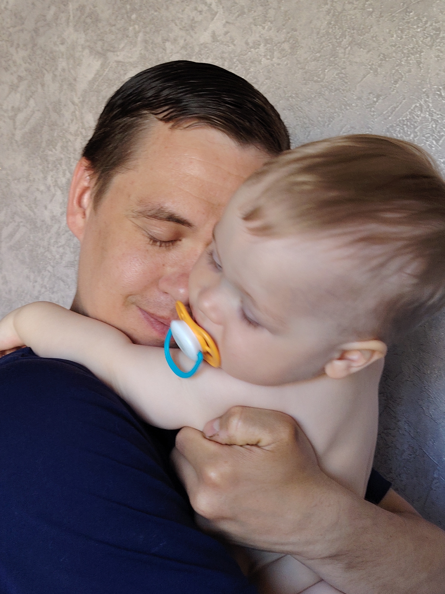 A medium shot depicts a man holding a baby. The man is wearing a dark blue t-shirt and has dark hair slicked back. He is looking down with his eyes closed and a slight smile on his face. The baby, who is fair-skinned and appears to have light brown hair, is snuggled against the man's chest and is using a pacifier. The pacifier is orange and white, with a blue handle. The baby's arm is wrapped around the man's neck. The background is a light gray wall with a textured surface. Based on context, this is likely in Chaikovsky, Russia.