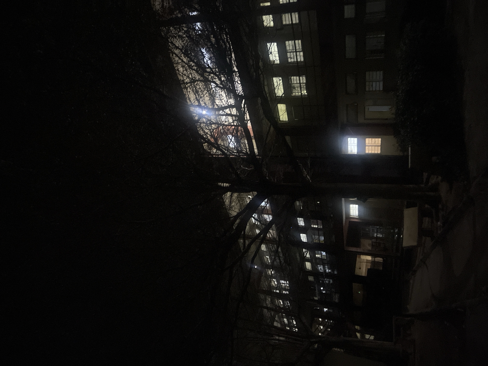 The image depicts an urban scene at night. A building with multiple illuminated windows dominates the background. A tree with bare branches is silhouetted in the foreground, partially obscuring the building. The environment is dark, with limited ambient light. The location cannot be determined to be Minna, Nigeria from the provided image.