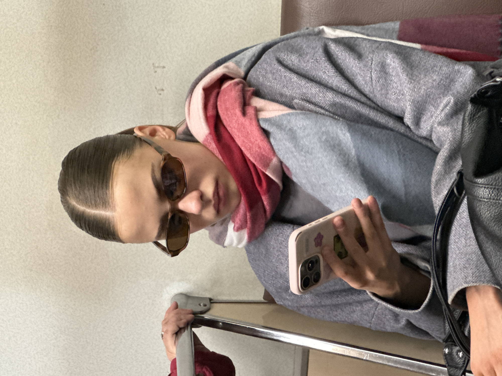 A young woman with slicked-back hair and sunglasses sits indoors, possibly on public transport. She wears a grey coat and a pink and grey scarf. She holds a pink smartphone with a floral and avocado sticker on the back. Her left hand is on a metal bar. A black leather bag is visible on her right. The background includes a beige wall and a brown seat. There is no information to confirm the location is Khimki, Russia.