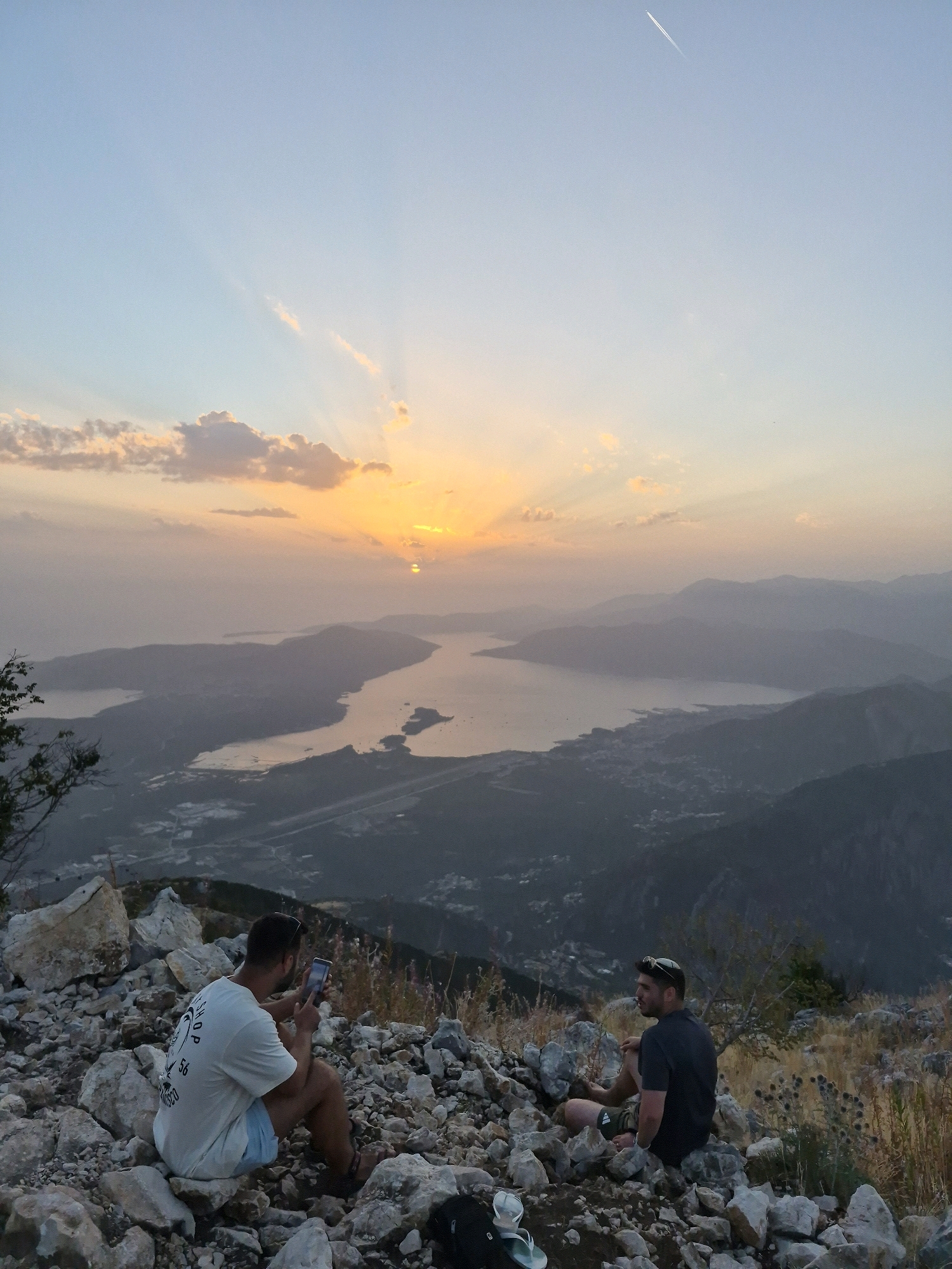 The image depicts a panoramic view of a bay, possibly the Bay of Kotor, near Kuk, Montenegro, during sunset. Two men are seated on a rocky outcrop in the foreground, facing the scenic vista. The man on the left, wearing a white t-shirt and light blue shorts, is holding a smartphone and appears to be taking a photograph. The man on the right, dressed in a dark t-shirt and dark shorts, is looking out at the landscape.
The sky is illuminated by the setting sun, casting warm hues across the clouds and creating visible sunbeams. Distant mountains form layers of silhouettes against the hazy horizon. Below, a coastal town or village is visible nestled along the edge of the bay, with lights beginning to appear as dusk settles. The terrain in the foreground is rough and rocky, with sparse dry vegetation. The overall atmosphere is serene and contemplative, capturing a moment of appreciation for the natural beauty of the location.