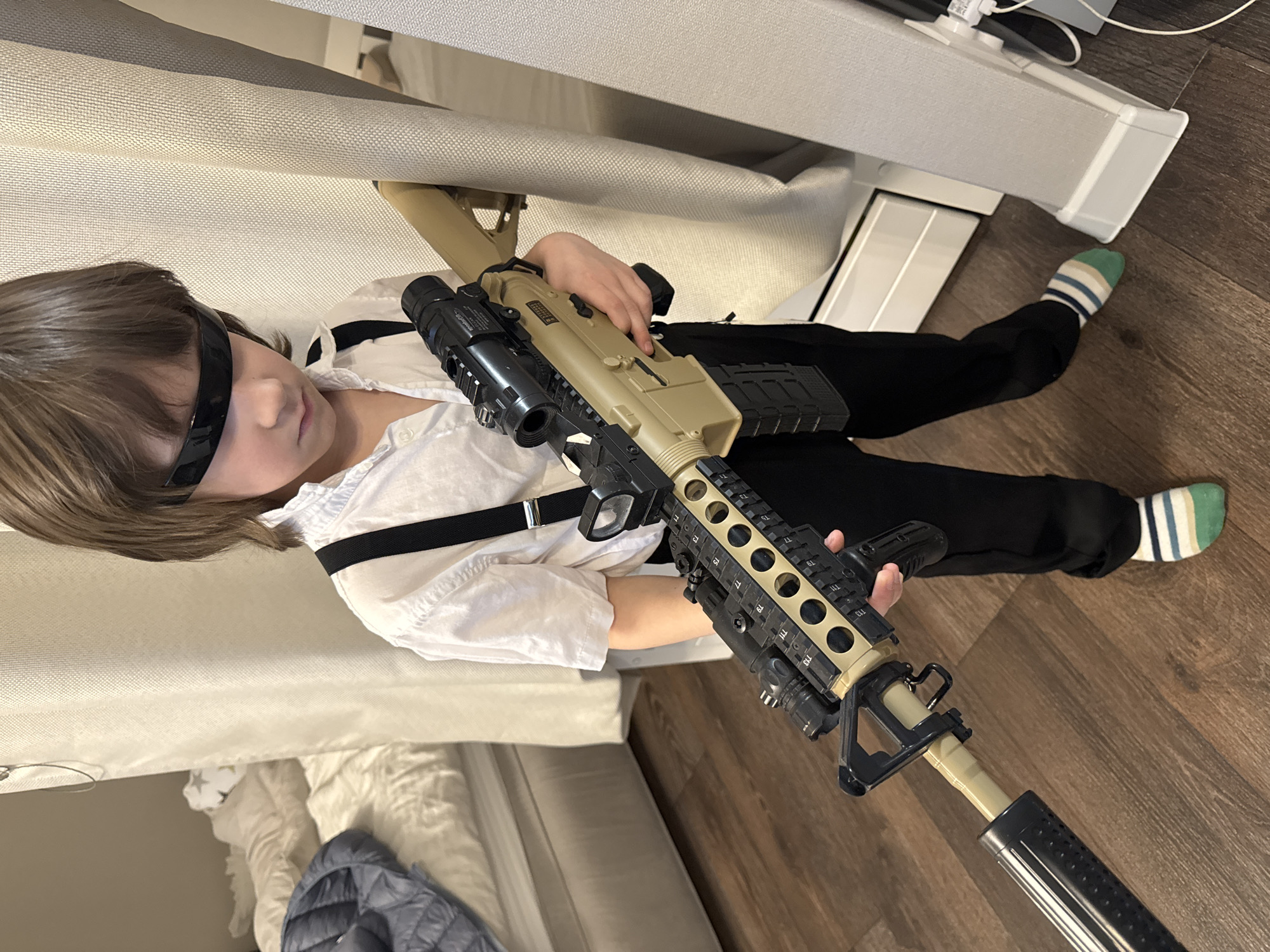 A child wearing black glasses, a white shirt, and black suspenders is lying on a floor made of wooden planks. The child is holding a large, beige and black toy assault rifle. They are wearing black pants and socks with green, white, and blue stripes. The child is positioned near a white couch and a white object that could be a piece of furniture or equipment. The setting appears to be indoors, possibly in a home. The city of Chaikovsky, Russia, is not visible in the image.
