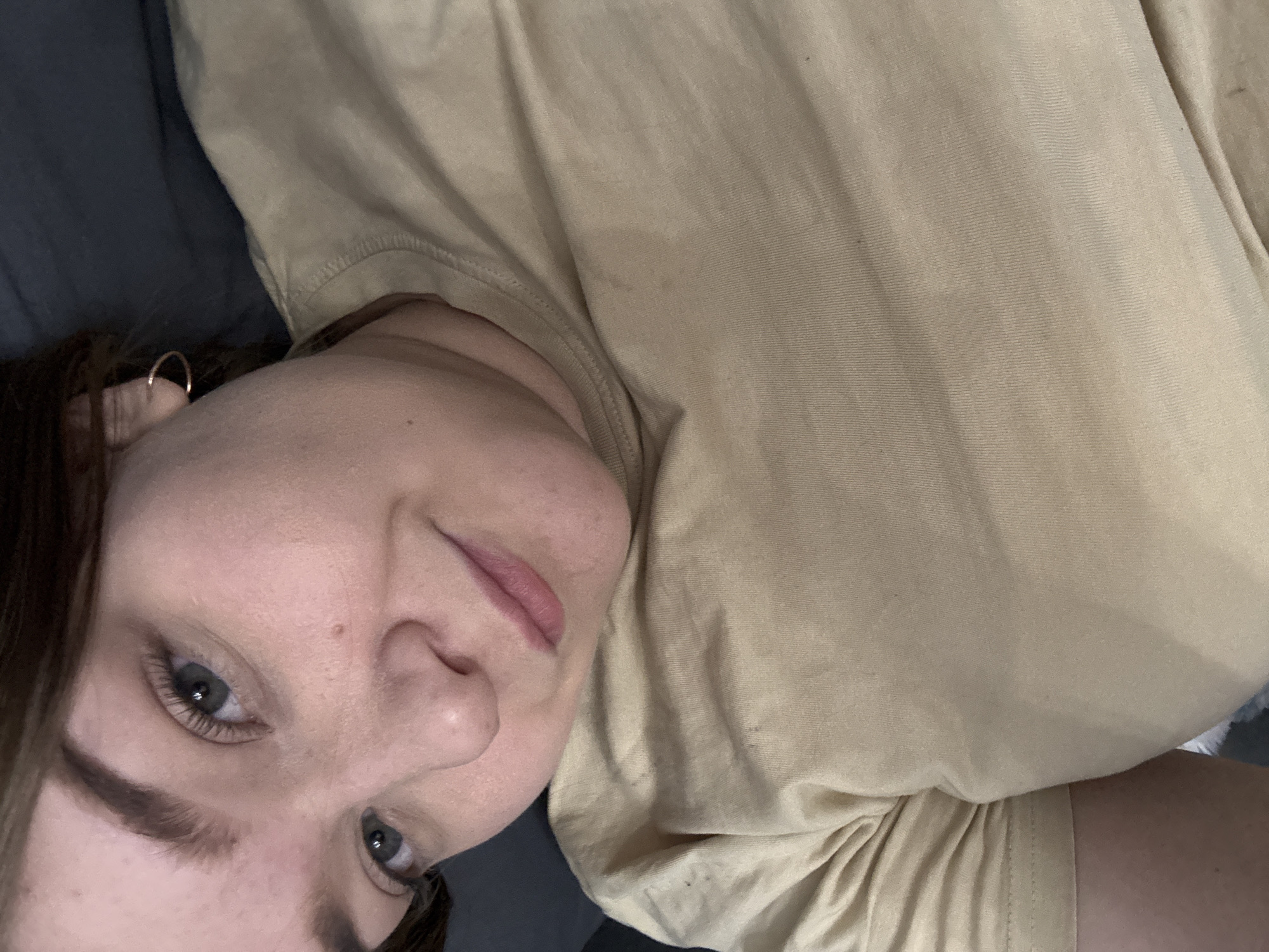 A woman with fair skin and blue-gray eyes is depicted in a close-up, slightly angled view. She has dark brown hair and is wearing a tan-colored t-shirt. The background includes a dark gray or black surface, possibly part of a sofa or bed. The lighting appears to be soft and even. There is no information about the location in the city of Chaikovsky, Russia.