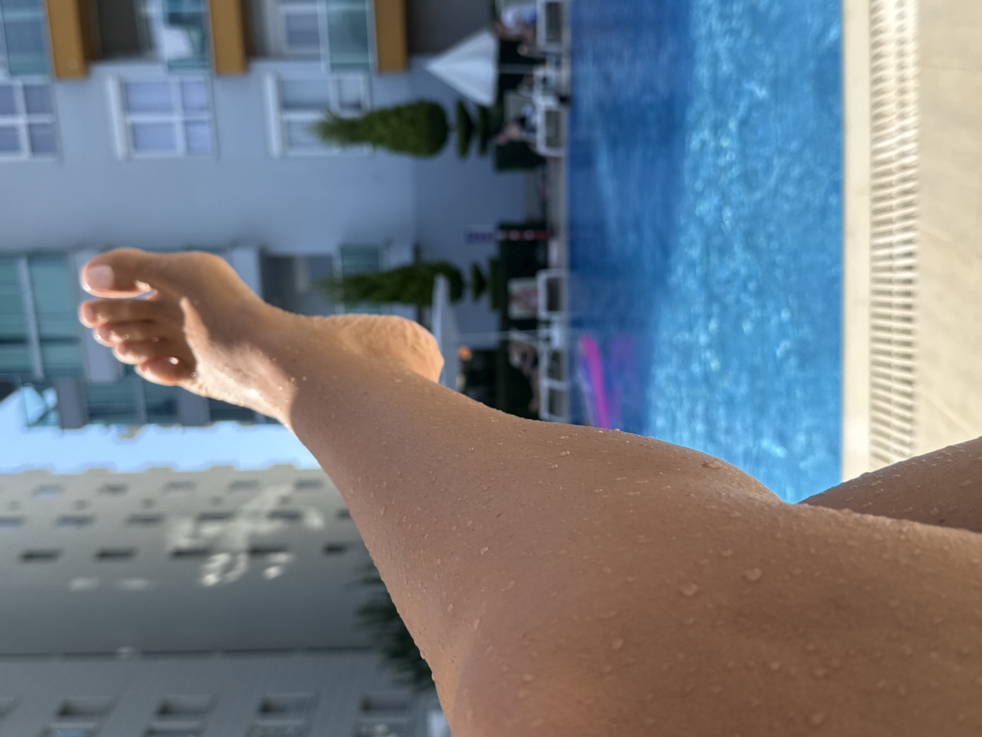 The image shows a close-up view of a person's wet leg and foot, extending into the frame from the lower left. The skin is covered in small water droplets, indicating recent exposure to water. The leg is bent at the knee, with the foot pointed upwards and slightly towards the right.
In the background, a bright blue swimming pool dominates the lower half of the image, with ripples and light reflecting on its surface. The edge of the pool is visible, with a white grated drain cover. Beyond the pool, a resort area is suggested by lounge chairs and umbrellas, with people faintly visible relaxing.
Behind the resort area, two modern, multi-story buildings are visible against a clear blue sky. The buildings have a light gray facade with numerous windows and balconies. One building features orange accents on some balconies. The overall impression is a sunny day at a resort in Alanya, Turkey, likely at a hotel or apartment complex with a swimming pool.