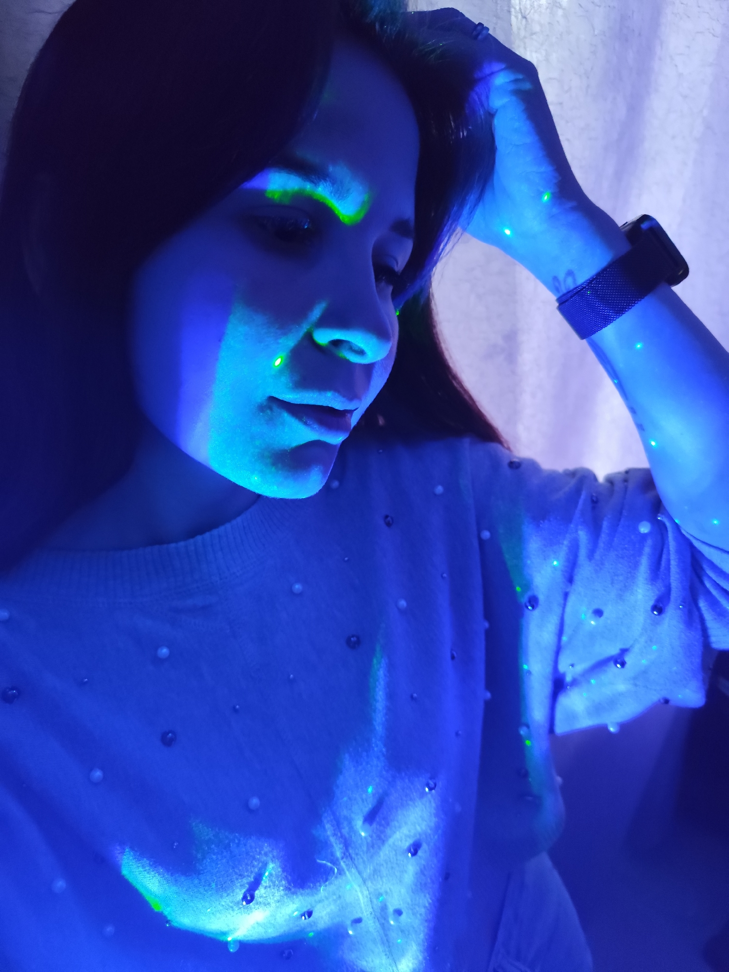A woman is illuminated by blue and green light in an indoor setting. She is wearing a light-colored sweater with embellishments. Her left hand is raised near her head, and a watch is visible on her wrist. The background appears to be a textured white curtain or wall. There is no evidence to suggest this image was taken in Фоки, Russia.
