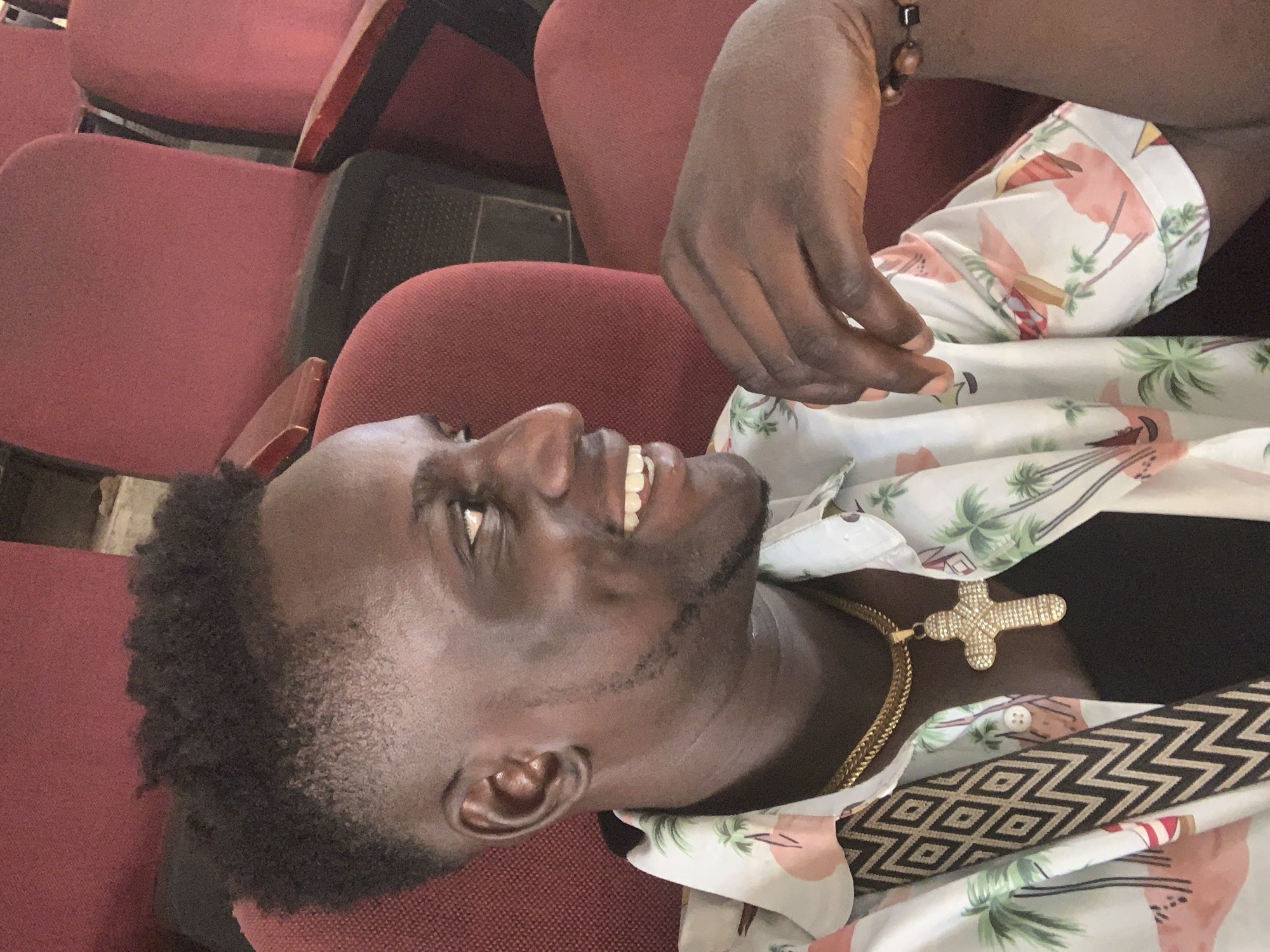 The image shows a young Black man with dark skin, short, curly hair, and a beard. He is wearing a white short-sleeved shirt with a tropical print featuring pink flamingos and palm trees, a gold chain, and a gold cross pendant. The man is smiling widely, with his eyes closed and his head tilted back, suggesting he is experiencing joy or amusement.
He is seated in a red theater or auditorium seat. The background is blurred but appears to be a row of similar red seats, indicating an indoor setting like a cinema or lecture hall. The lighting appears to be artificial.
A black bracelet with brown beads is visible on his left wrist. The overall impression is a candid shot, capturing a moment of happiness for the subject. The image orientation is landscape, with the man's head towards the right side of the frame.