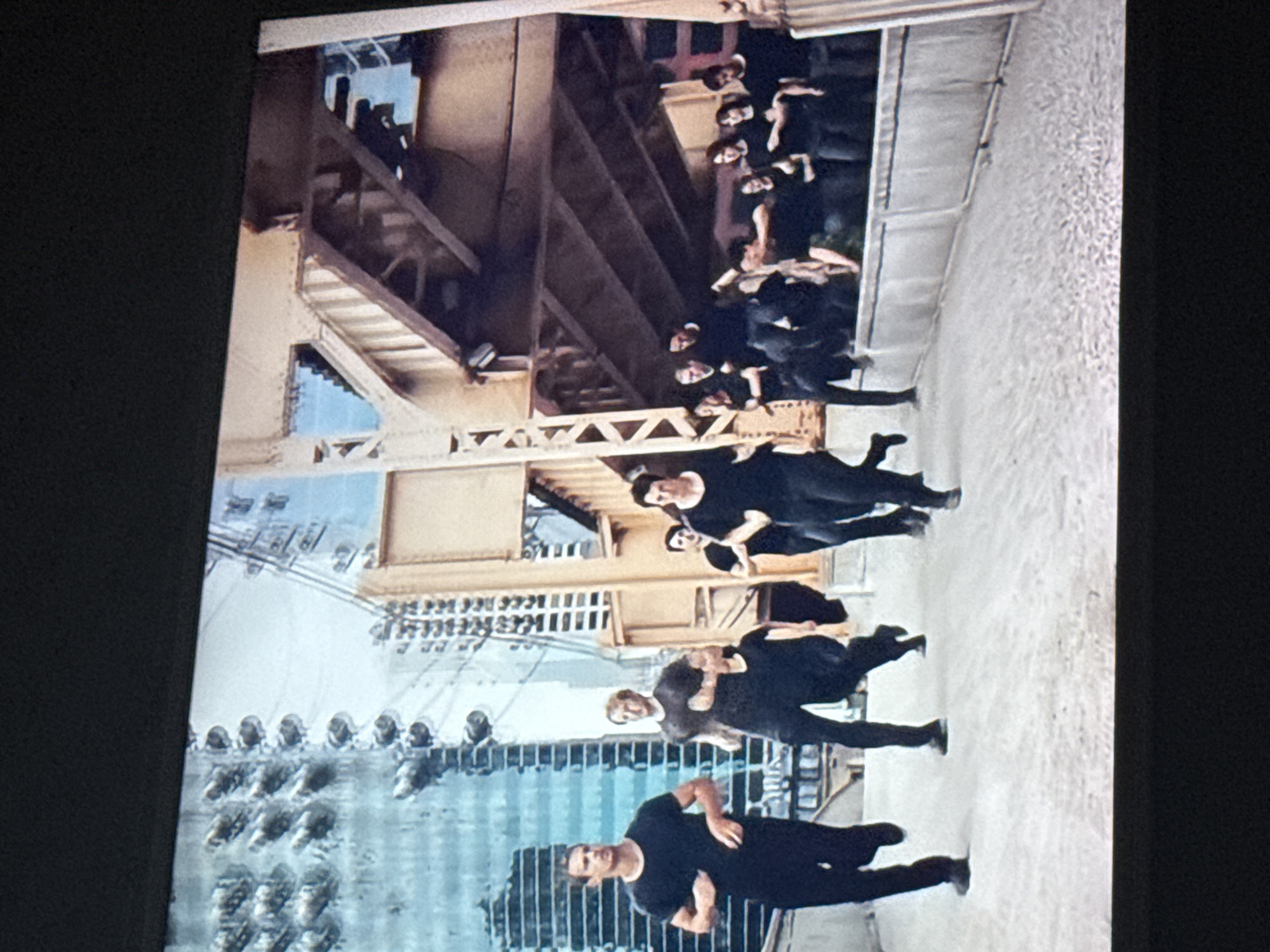The image depicts a group of people dressed in black performing a dance or movement sequence in an urban setting. A yellow-painted bridge structure is overhead, and a modern building with numerous round, protruding architectural details appears in the background. The location is likely in a city, potentially Mytishchi, Russia, given the prompt's reference. The dancers are positioned on a light-colored surface, possibly a sidewalk or pavement. They are spaced out in a line. The image suggests a performance art piece or a rehearsal taking place in a public urban space.