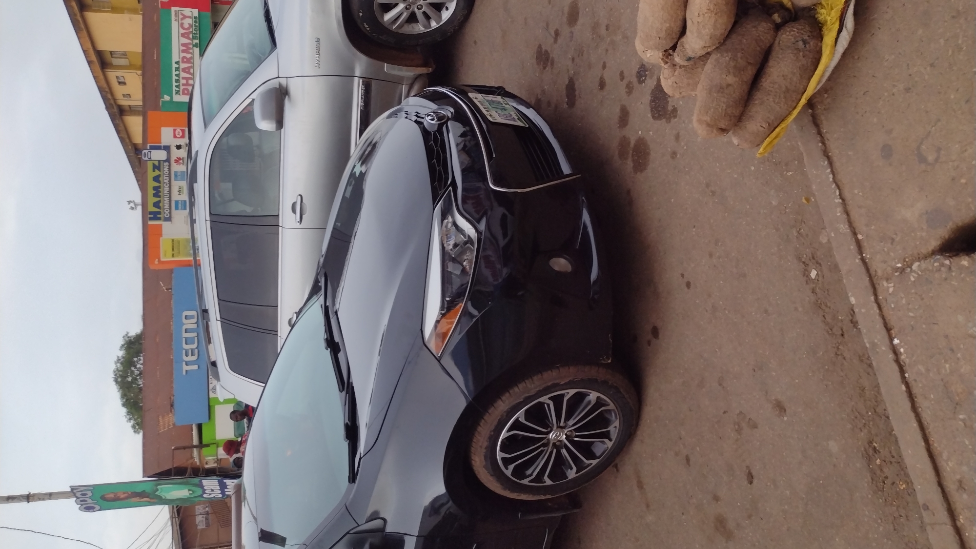 The image presents a street scene in Jos, Nigeria. Dominating the view are two vehicles: a black car in the foreground and a silver SUV behind it. In the upper right corner, a pile of yams rests on what appears to be a yellow cloth or container. Behind the vehicles, various business signs are visible, including "Nasara Pharmacy & Stores", "Hamaz Communications", and "Tecno". An "Opay" sign on a pole is situated on the left side of the image. The street surface appears to be made of concrete, and there is an indistinct line of curb. People are partially visible behind the Tecno sign, suggesting some level of pedestrian activity.