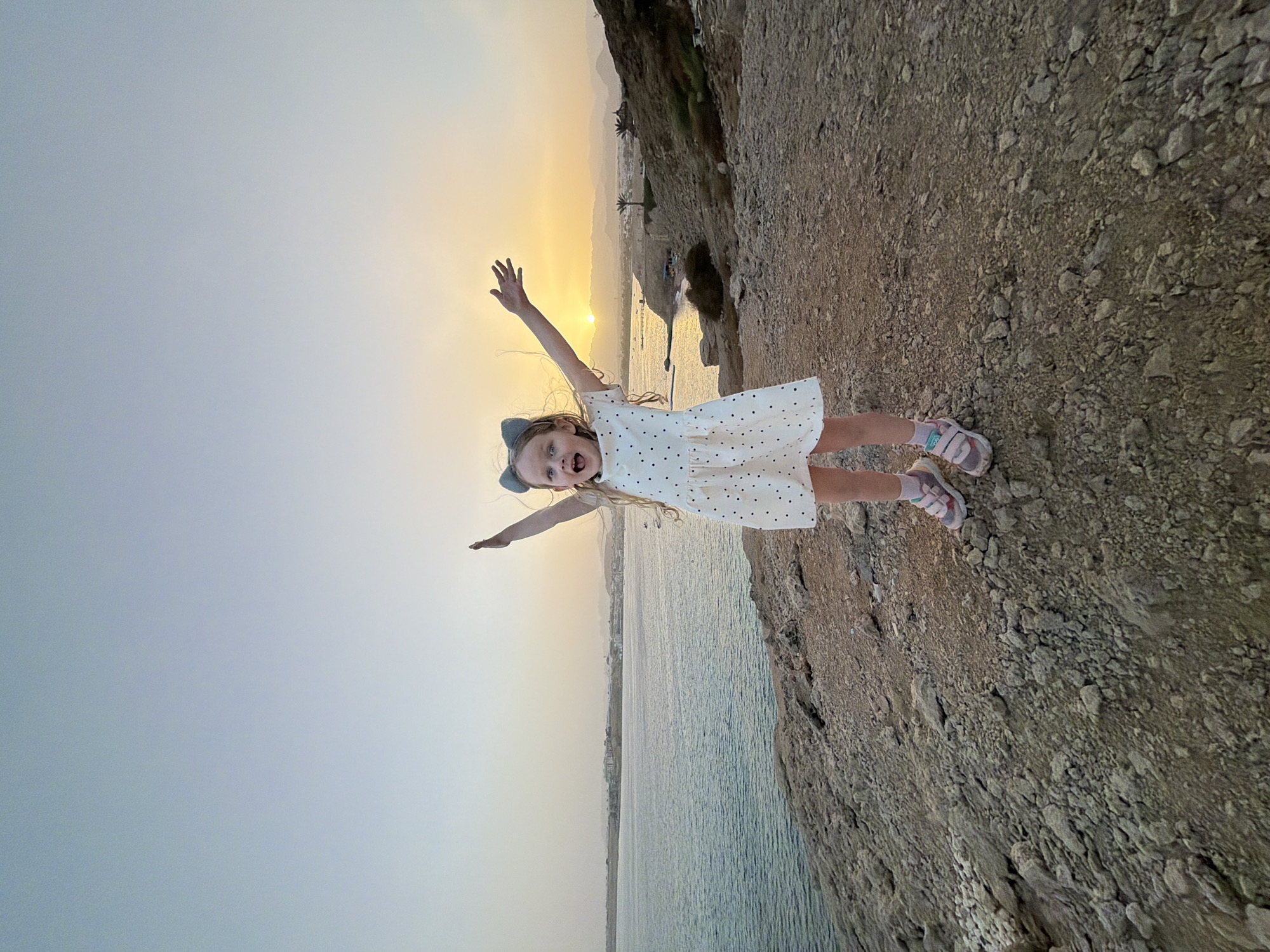 A child stands on a rocky outcrop with her arms outstretched, appearing to be flying. She is wearing a white dress with black polka dots and pink sandals. The backdrop features a body of water and the city line of Sharm El Sheikh, Egypt, with the sun setting in the background. The sky is pale blue near the horizon, blending into a hazy yellow around the sun.