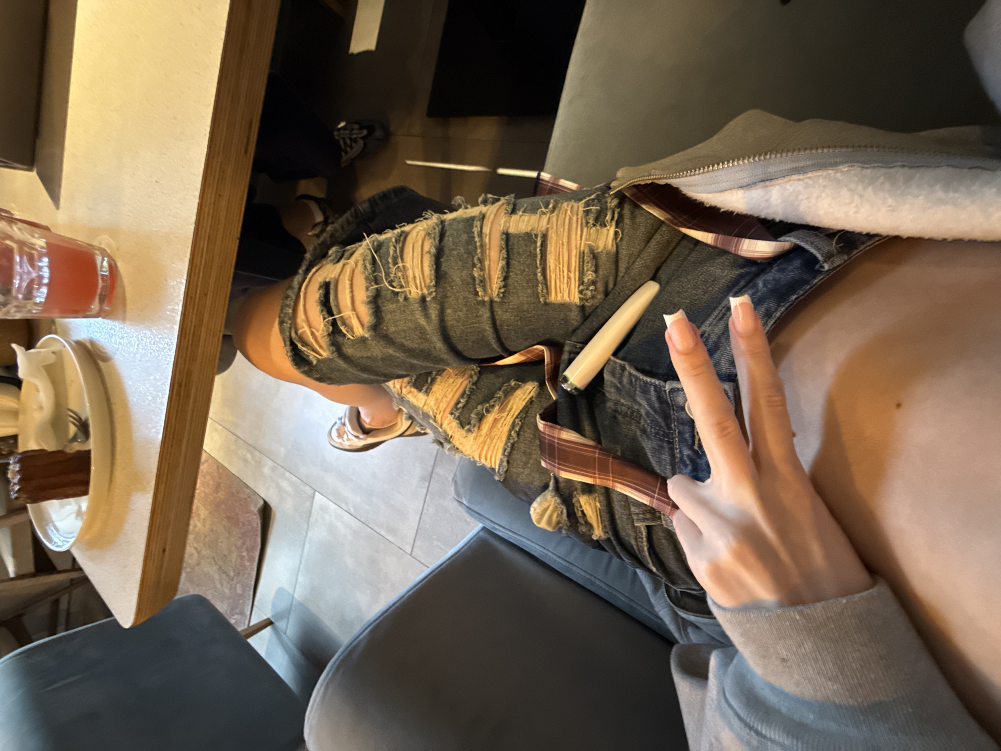 This media file shows a close-up, vertically oriented shot of a person sitting in what appears to be a cafe or restaurant in Khimki, Russia. The primary subjects are the person's legs, wearing distressed denim shorts with visible patches of plaid fabric underneath. A light-colored object, possibly a vape pen or e-cigarette, rests on the shorts. The person's right hand is in the frame, making a peace sign gesture with painted fingernails. They are wearing a grey hooded sweatshirt.
The setting includes a light-colored table with a glass containing a red beverage and a plate with a piece of chocolate cake on it. A dark-colored chair is visible in the foreground. The flooring appears to be tiled.
The image is likely taken during daylight hours, given the lighting conditions. There are no explicit indicators of the weather. The overall mood is casual and relaxed, suggested by the clothing, the setting, and the hand gesture. No visible text is present in the image.