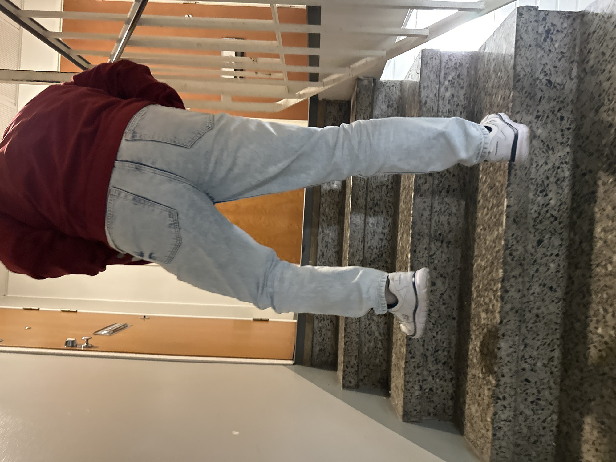 A person is walking up a concrete staircase inside a building. The person is wearing a red jacket and light-wash jeans, and white athletic shoes. The staircase has a metal railing on the right side, and a wooden door is visible on the left. The walls are light-colored.
