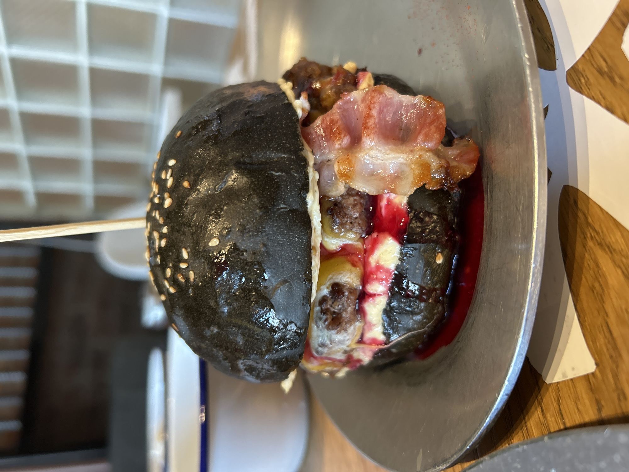 A close-up, eye-level shot captures a gourmet burger with a black bun, served on a grey metal plate. The burger is a towering creation featuring a thick, juicy beef patty, melted yellow cheese, a fried egg, and crispy bacon strips. A vibrant red sauce oozes from the sides of the burger, pooling slightly on the plate. The bun is generously sprinkled with sesame seeds, and a wooden skewer pierces the top bun, holding the burger together. The background is softly blurred, showing hints of a modern interior with a textured brick wall and white wall accents. The lighting is bright, highlighting the textures and colors of the food.