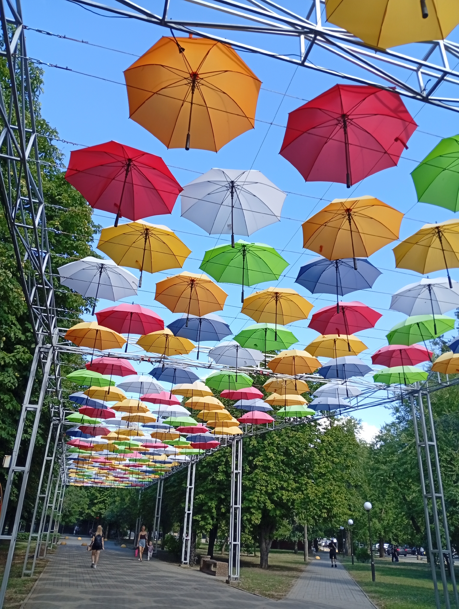 The image captures a public walkway in the city of Pokrov, Ukraine, adorned with a vibrant canopy of colorful umbrellas suspended overhead. The umbrellas, in shades of yellow, red, green, blue, and white, create a striking visual spectacle against the clear blue sky. The walkway is paved with gray interlocking stones and bordered by lush green trees on both sides. In the background, more trees and some parked cars can be seen. Several individuals are walking along the path. In the foreground, a young woman with blonde hair tied back, wearing a black dress, walks away from the camera. Further ahead, another woman, with dark hair, wearing a gray t-shirt and shorts, walks with a young child. Several lampposts are visible along the walkway. The overall scene depicts a lively and artfully decorated public space on a sunny day.