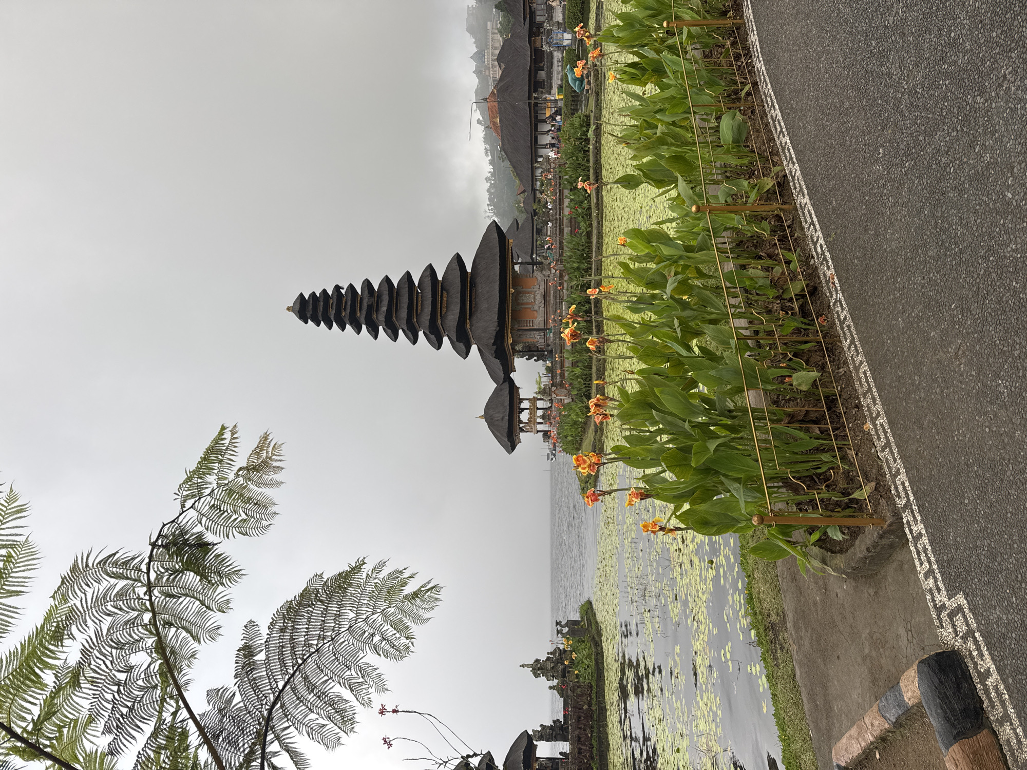 The image shows the Ulun Danu Beratan Temple in Bali, Indonesia, reflected in the calm waters of Lake Beratan. The temple's distinctive tiered pagoda-like structure is prominently featured, with the reflection creating a symmetrical visual effect. The surrounding landscape includes lush greenery and aquatic plants floating on the lake's surface. A paved pathway runs along the edge of the water, lined with a decorative fence and vibrant flowers. The sky is overcast, casting a soft, diffused light over the scene.