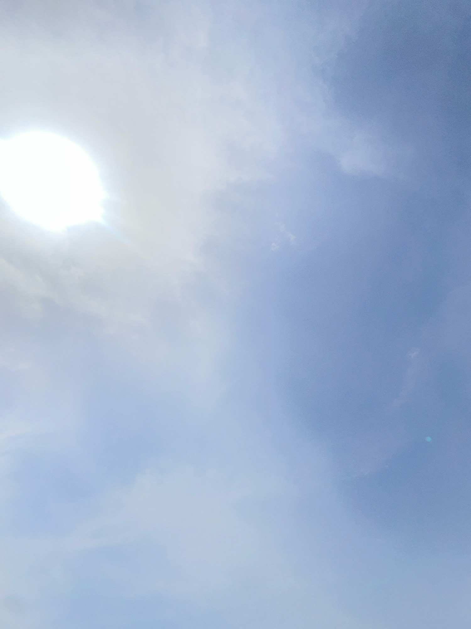 The image shows a bright sun shining in a partly cloudy sky. The sky is a pale blue with scattered white clouds, some of which are thin and wispy, while others are more substantial. The sun is located in the upper left quadrant of the image and is overexposed, appearing as a bright white disc. There is a slight lens flare in the lower right quadrant of the image. The scene appears to be outdoors, likely in La Herradura, Spain, given the context. There are no people, subjects, or objects visible in the image other than the sky and the sun.