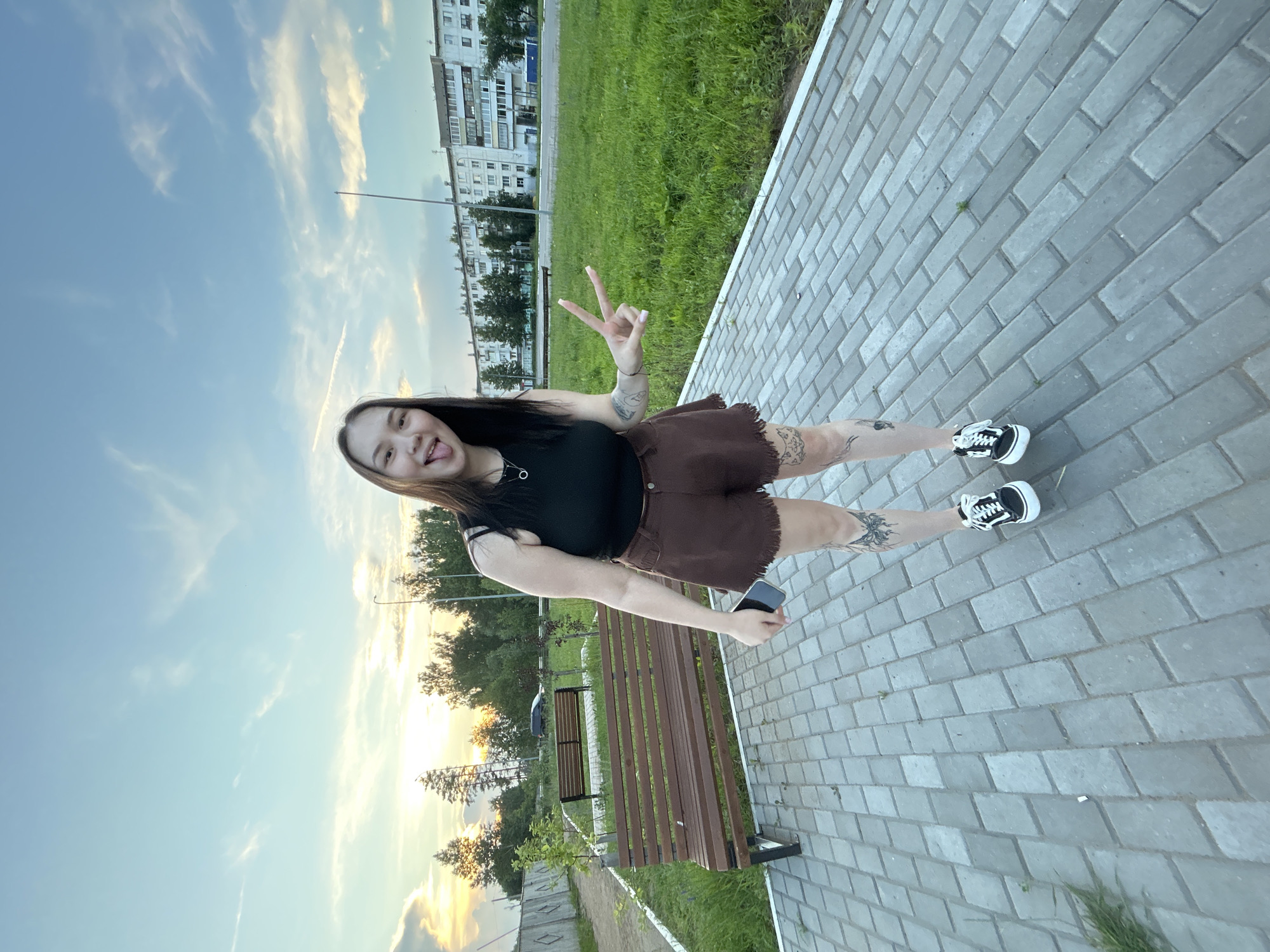 A young woman is posing for a photo in a public park in Chaikovsky, Russia. She is wearing a black tank top, brown shorts, and black sneakers. She has tattoos on her legs and is sticking her tongue out at the camera, holding up a peace sign with her right hand. She is standing on a paved walkway, with a wooden bench to her left and a grassy area behind her. In the background, there are trees, a fence, and apartment buildings. The sky is blue with some clouds, and the sun is setting, casting a warm glow over the scene.