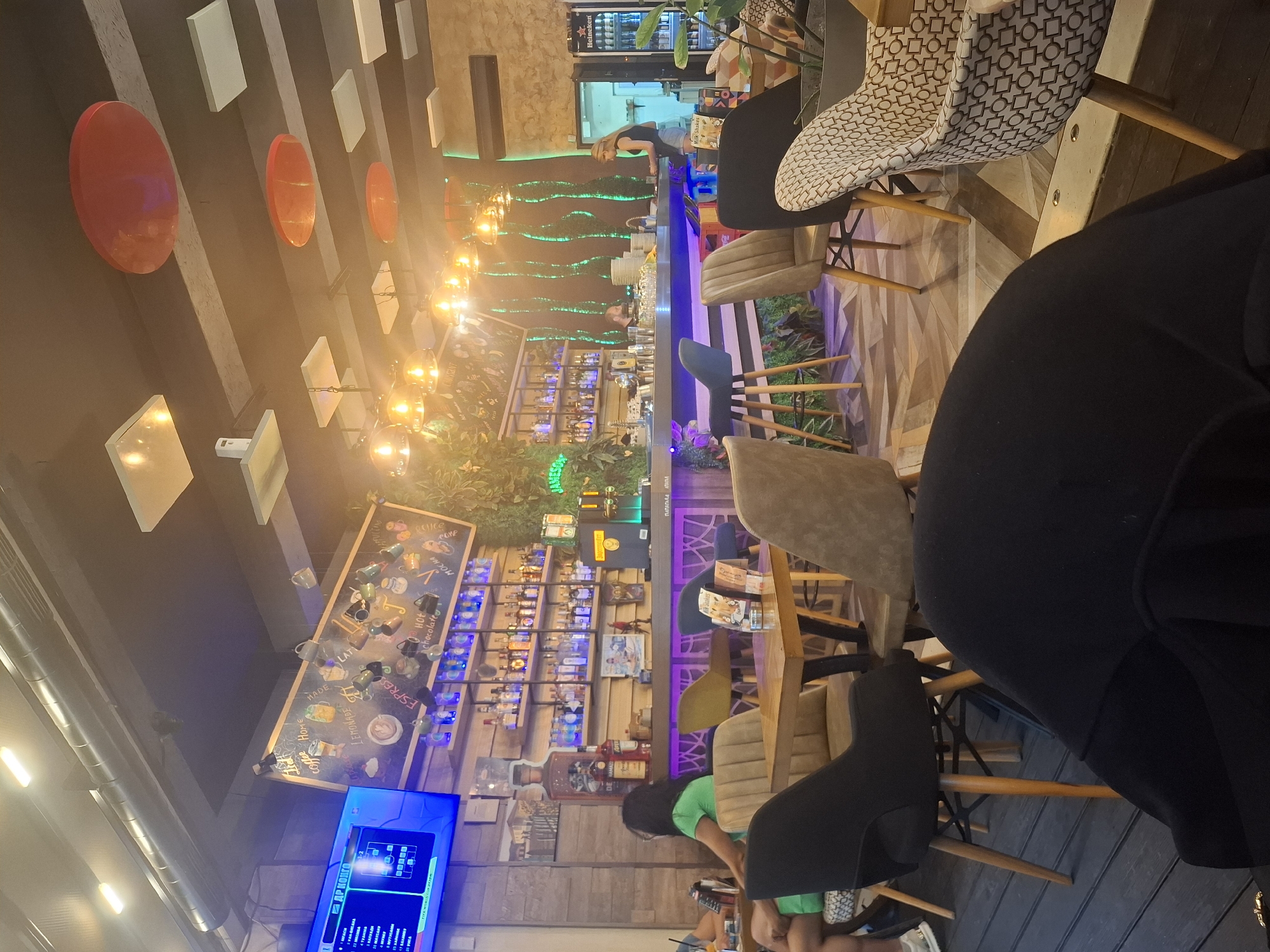 This media file captures an indoor scene, likely a cafe or bar, with a dimly lit and cozy ambiance. The perspective is angled, giving a sense of immersion. Several individuals are present, though their faces are not clearly visible, suggesting a casual or everyday setting.
In the foreground, the back of a person in a black outfit is prominently featured, along with a chair. The midground reveals a bar area with shelves stocked with bottles, illuminated by colorful lights, implying it is operational. A woman is visible behind the counter, seemingly engaged in work. The scene is further populated by various chairs and tables, some with patterned upholstery, indicating customer seating.
A notable object is a large chalkboard-style menu displayed on the wall, featuring handwritten text and drawings related to coffee and other beverages. Visible text includes "coffee," "LEMONADE," "ESPRES," "Hot chocolate," and "MOCHA." Other text includes "VAN-BUGARA," "КАЛКАЛИ," and "ДР КОНГО." A large television screen is also visible, displaying what appears to be a menu or information board.
The overall impression is that of a lively establishment, possibly during the evening or at a time when artificial lighting is dominant. The environment seems relaxed and inviting, with a focus on drinks and social gathering. The eclectic decor, including what looks like some greenery and patterned elements, adds to the character of the space.