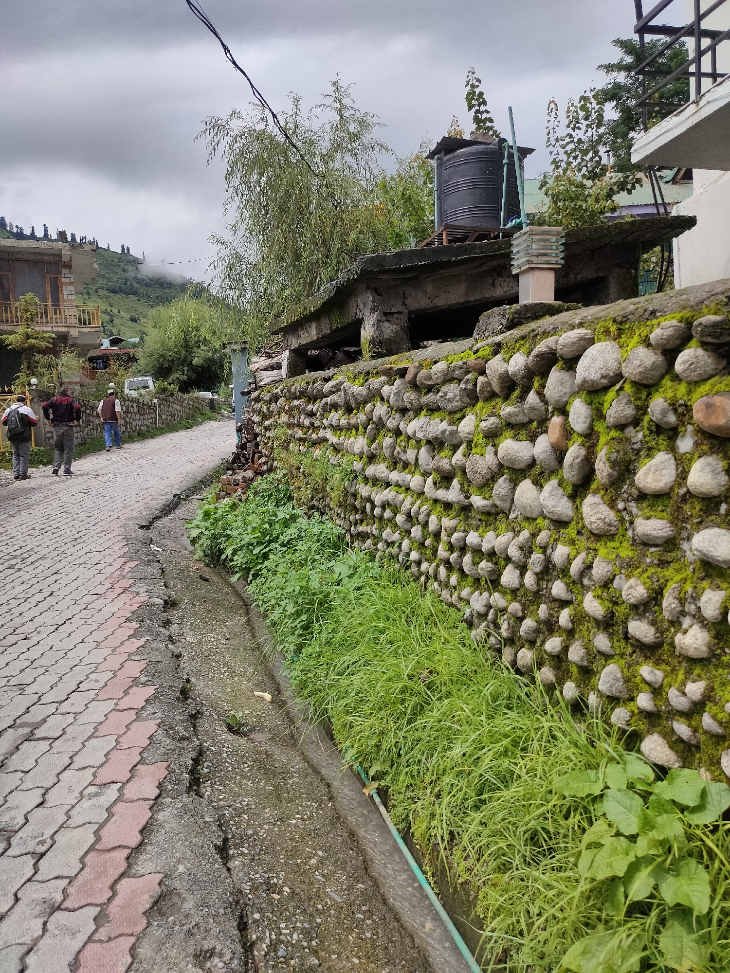 In Burwa, India, a paved road with interlocking concrete pavers, some colored red, runs from the foreground up a gentle incline. To the left, three individuals walk away from the viewer. The first person, on the left, wears a light-colored backpack. The second person, in the middle, is dressed in a maroon jacket and dark pants. The third person, on the far right, wears a dark vest over a lighter shirt and dark pants. A white van is parked further up the road on the left. Adjacent to the road, a narrow, shallow channel carries flowing water. On the right side of the road, a roughly constructed stone wall, covered in moss and lichen, forms a boundary. Above the stone wall, there is a concrete structure with a black water tank and what appears to be a light fixture. Behind this, a hillside with trees and some buildings is visible, shrouded in mist under an overcast sky.