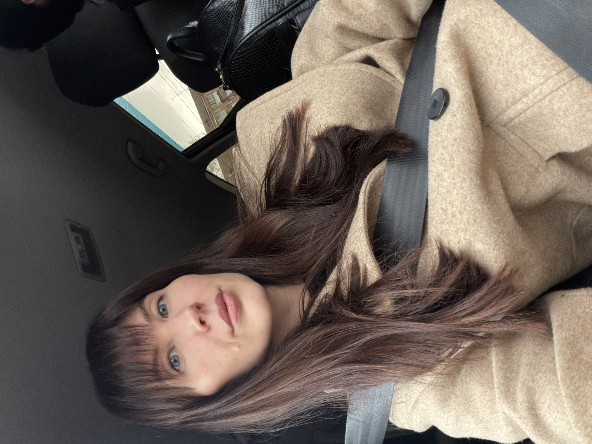A young woman with fair skin and long brown hair sits in the driver's seat of a car, wearing a light tan wool coat and a seatbelt. The car has a sunroof, visible behind her, with a glimpse of overcast sky and an unidentified building exterior. The car's interior is dark gray. Based on visual cues like the architectural style of the building, the image was likely taken in Perm, Russia.