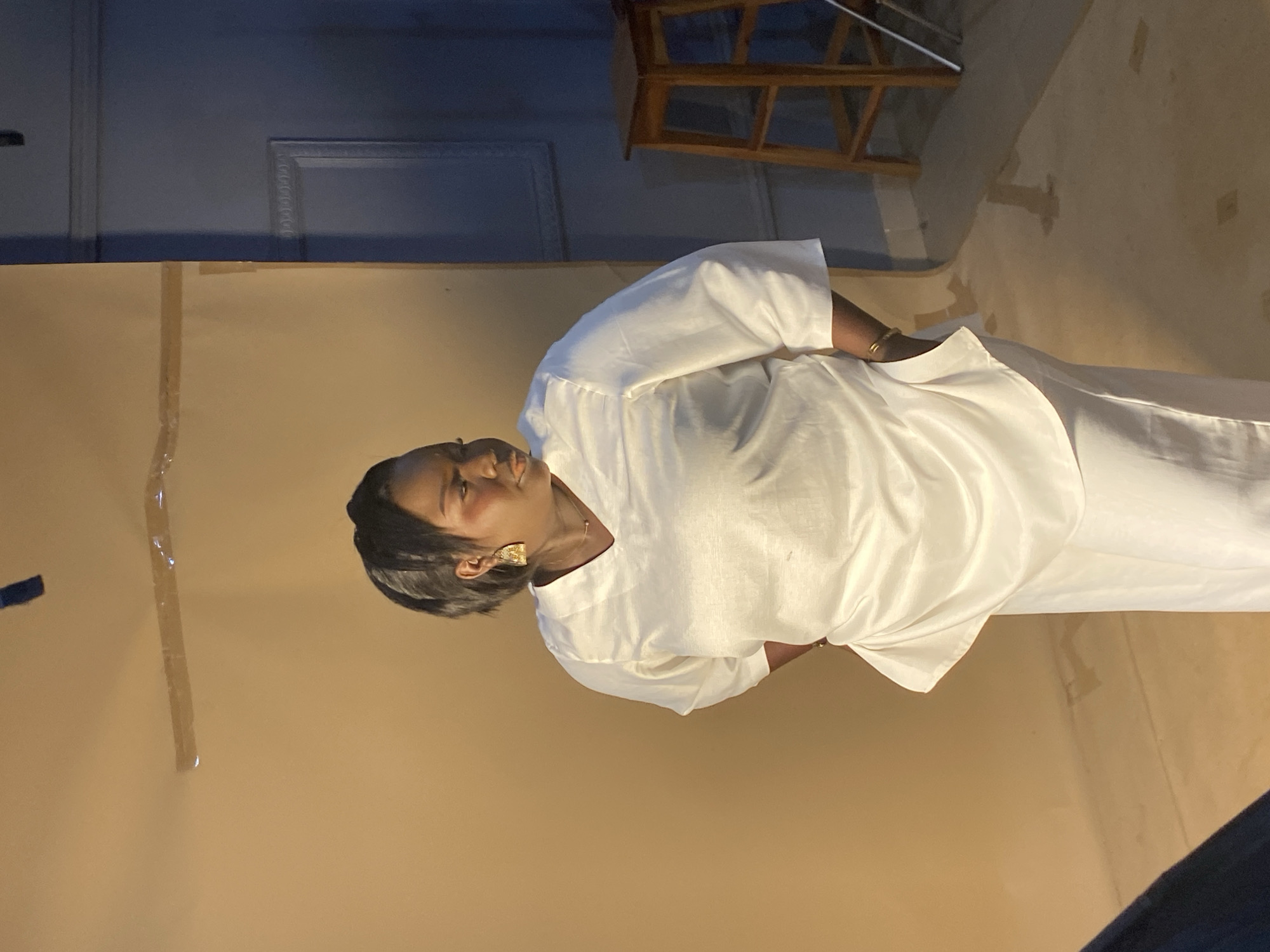 The media file shows a medium shot of a Black woman standing in what appears to be a studio setting, with a tan backdrop. She is wearing a white, loose-fitting top and white pants. Her hair is black and styled in a bob. She is wearing large, gold, geometric earrings and a thin gold bracelet on her right wrist. Her head is tilted to her left, and she is looking upwards and to her left. Her facial expression is neutral, with her mouth slightly open. The lighting suggests an indoor environment, possibly with artificial light. The background is a plain, light brown color, with some subtle texture or variations in shade. In the upper left portion of the image, a glimpse of a dark blue wall with decorative molding is visible, along with a dark wooden stool or chair. The overall impression is that of a posed photograph, possibly for a portrait or a fashion shoot. The location context provided is Bukuru, Nigeria, though no specific cues in the image definitively indicate this location. There is no visible text in the image.