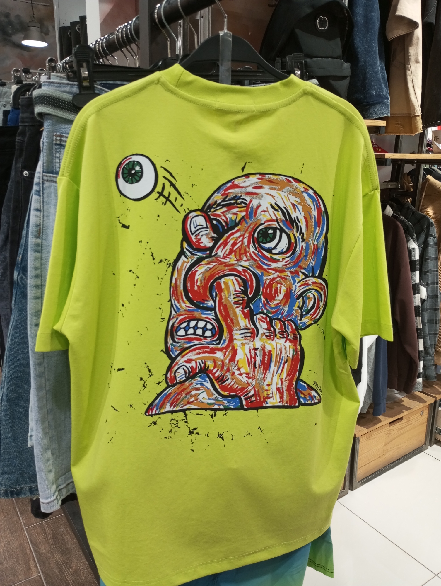 A bright green t-shirt hangs on a rack in a clothing store, showcasing a distorted, colorful graphic of a face with an eyeball hovering beside it. The shirt is surrounded by other garments, including jeans and various jackets, suggesting a retail environment. The clothing rack is part of a larger display, with wooden shelving and other merchandise visible in the background. The store appears well-lit, and the overall setting indicates a contemporary shopping space. The location, identified as Izhevsk, Russia, is based on external information not explicitly depicted in the image.