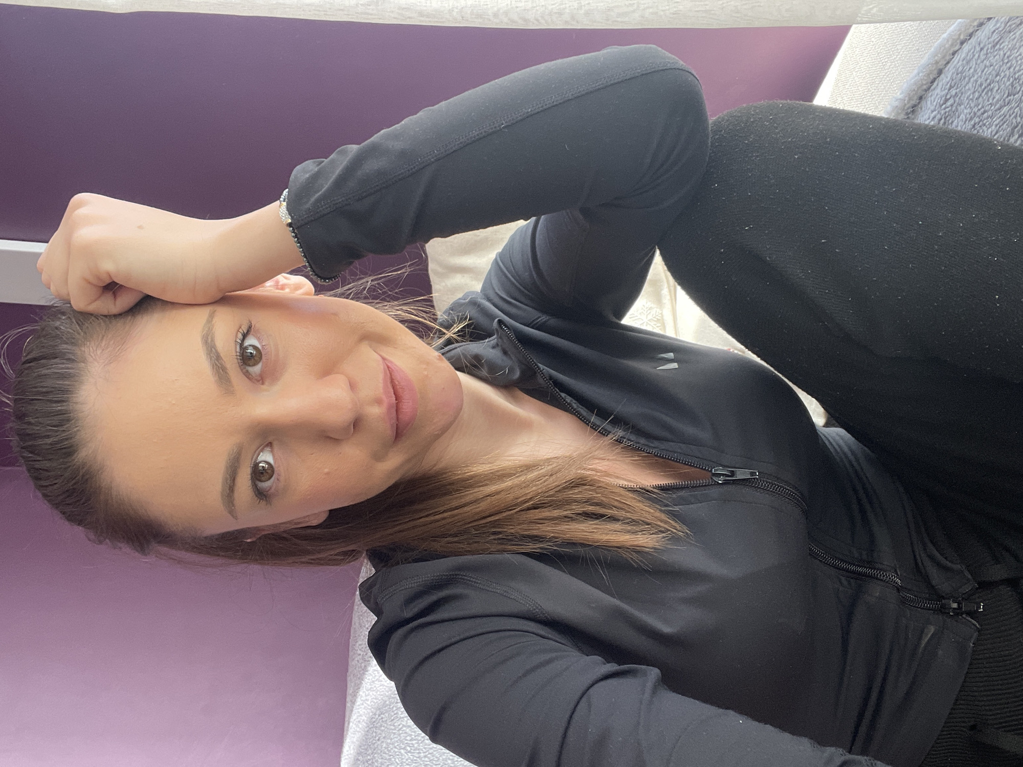 A woman is lying down. She has her hand on her forehead. She is wearing a black zip-up jacket and black leggings. The background appears to be a wall painted purple and a couch. It is not possible to determine the location within Sofia, Bulgaria, from this image.