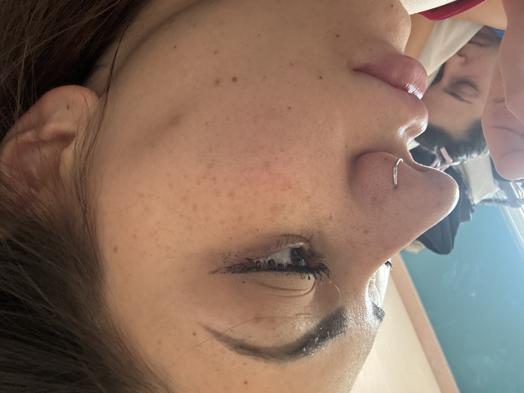 This media file is a close-up portrait of a person, likely female, taken in Sofia, Bulgaria, and presented on its side. The main subject's face is shown in profile, with her head oriented towards the right side of the frame, looking upwards from the viewer's perspective.
The subject has long, dark brown hair that partially frames her ear and falls against her cheek. Her skin is visible with numerous small, light brown freckles scattered across her cheek, temple, and forehead area. Her lips are full and appear to be enhanced with a pinkish gloss or lipstick. A silver, circular hoop nose piercing is prominent on her right nostril. Her right eye is open, showcasing dark eyeliner on both the upper and lower lash lines, along with visible mascara on her eyelashes. Her eyebrow is neatly shaped and appears to be filled in with a dark product.
In the upper right portion of the image, a reflection is visible, suggesting a mirror or a highly reflective surface. This reflection shows another person, possibly the same individual, with dark hair and closed eyes, appearing to be lying down or resting. Parts of the reflected background include light blue/teal and off-white surfaces, suggesting an indoor setting.
The lighting is bright and appears to be natural daylight, creating a soft, even illumination. There is no visible text in the image. The scene suggests a personal moment captured in a detailed, intimate manner.