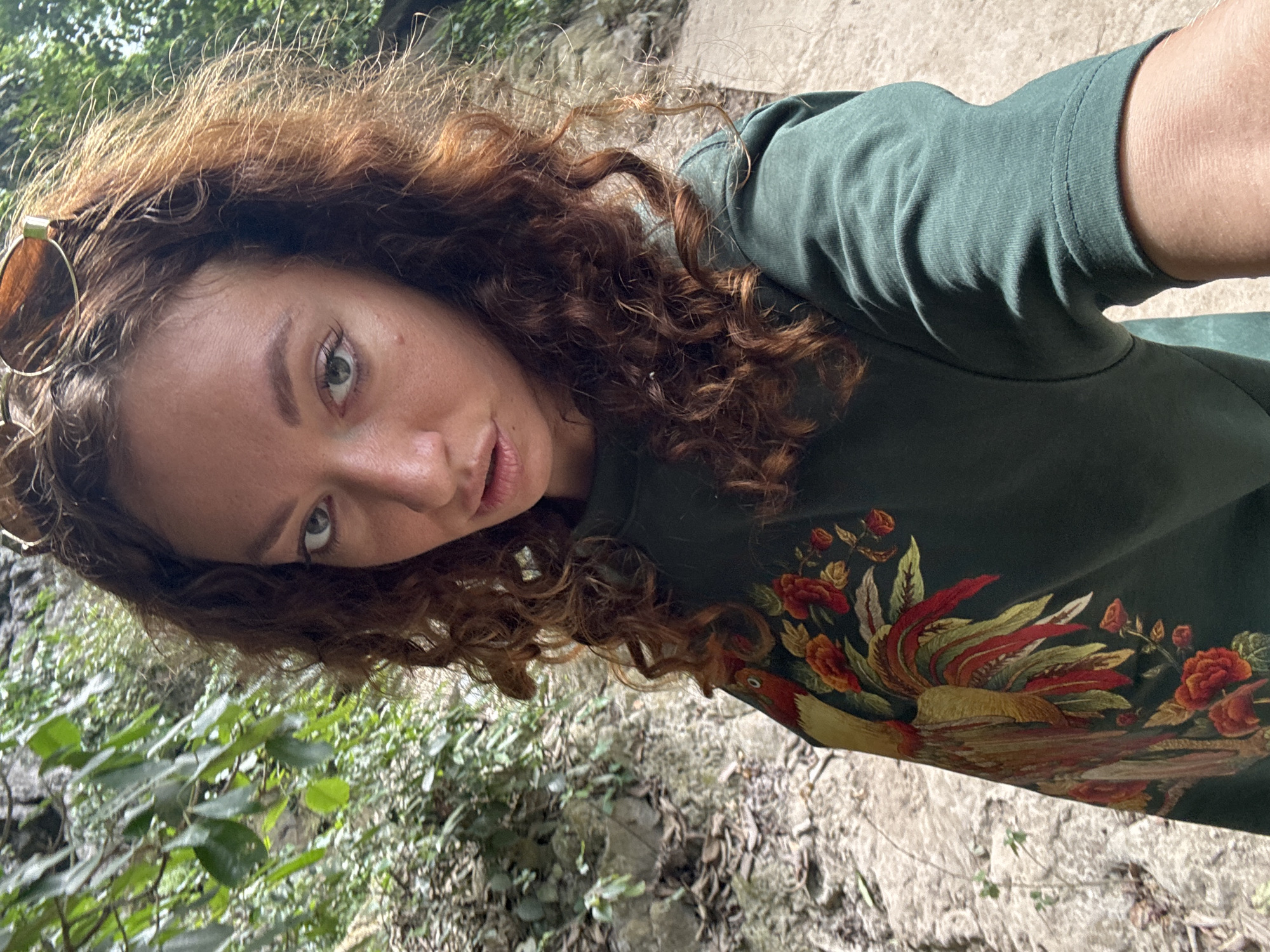 A woman with curly auburn hair, wearing sunglasses on her head and a dark green long-sleeved shirt with a rooster and floral embroidery, stands in a natural setting. She is looking at the camera with a neutral expression. The background consists of lush green foliage, rocks, and a dirt path, suggesting an outdoor, possibly rural or mountainous, location. The lighting indicates it is daytime.