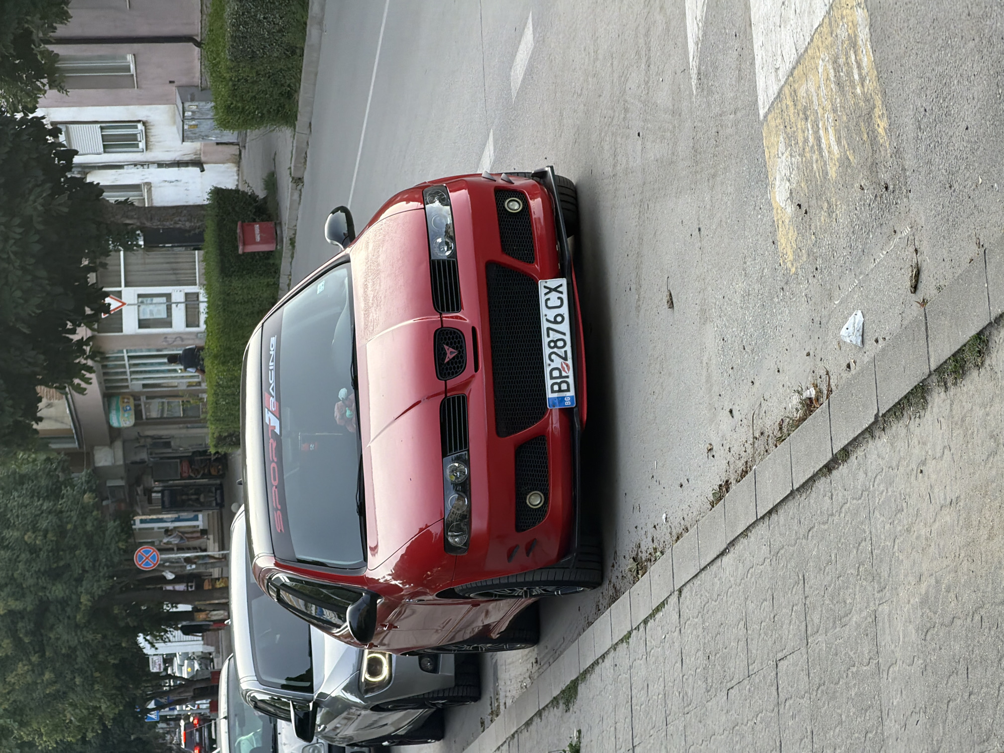 A red car, possibly a Seat Leon Cupra, is parked on a street in Gotse Delchev, Bulgaria. The car is the primary subject, with its front end facing towards the right of the frame. It has a sleek, sporty design with prominent headlights and a grille. A license plate reading "BP2876CX" is visible on the front bumper. The car is parked next to a sidewalk made of concrete pavers, and beyond that, a paved road. In the background, there are buildings with visible windows and balconies, some of which appear to be older structures. Green trees and hedges line the street, suggesting an urban or suburban setting. The lighting indicates it is daytime, likely late afternoon or early evening, given the soft shadows. No people are clearly visible in the immediate vicinity of the car. A "no parking" sign is visible in the distance, indicating traffic regulations. The overall scene is one of a parked vehicle on a street, with a backdrop of buildings and vegetation.