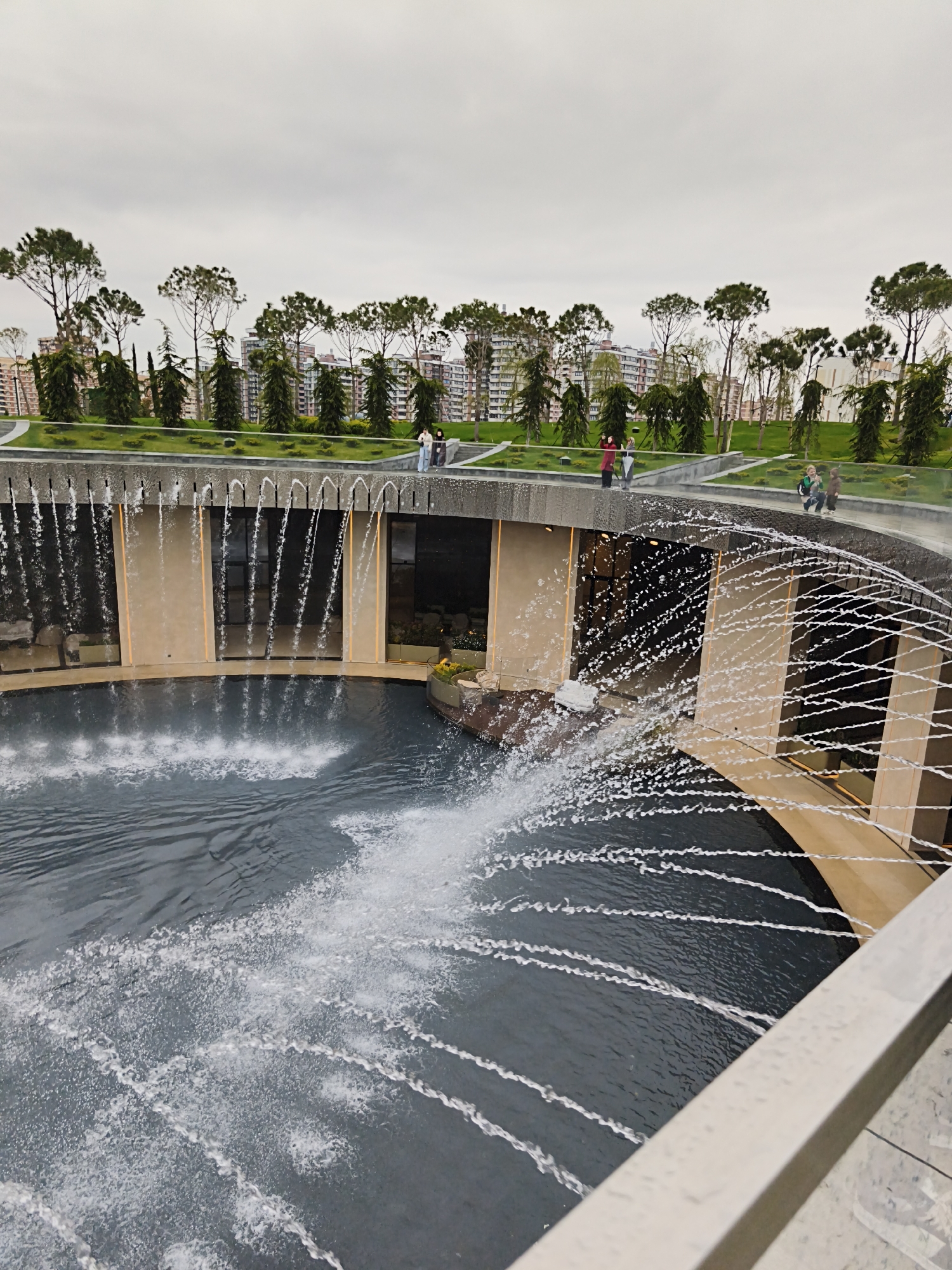 The image shows a large, circular fountain in the city of Krasnodar, Russia. The fountain is partially enclosed by a concrete structure with vertical openings. Water flows from the structure into the fountain. Several people are standing on the upper level of the concrete structure, appearing to be observing or taking pictures of the fountain. In the background, there is a row of trees and buildings under a cloudy sky.