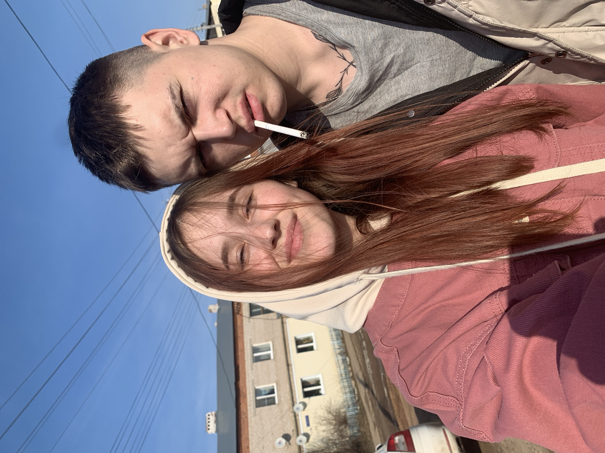 Two people pose closely for a selfie outdoors. The person on the left, presumably male, has short dark hair, closed eyes, and a cigarette in their mouth. They wear a gray t-shirt, a black jacket and a beige jacket on top. A tattoo of leaves is visible on their neck. The person on the right, presumably female, has long, reddish-brown hair partially obscuring their face. They are wearing a pink hooded jacket with a beige hood. Both individuals have their eyes closed and appear to be smiling. The background includes a clear blue sky, power lines, a multi-story building with windows, and a glimpse of a white car. Based on the building architecture visible in the image, the location is in the city of Фоки, Russia.