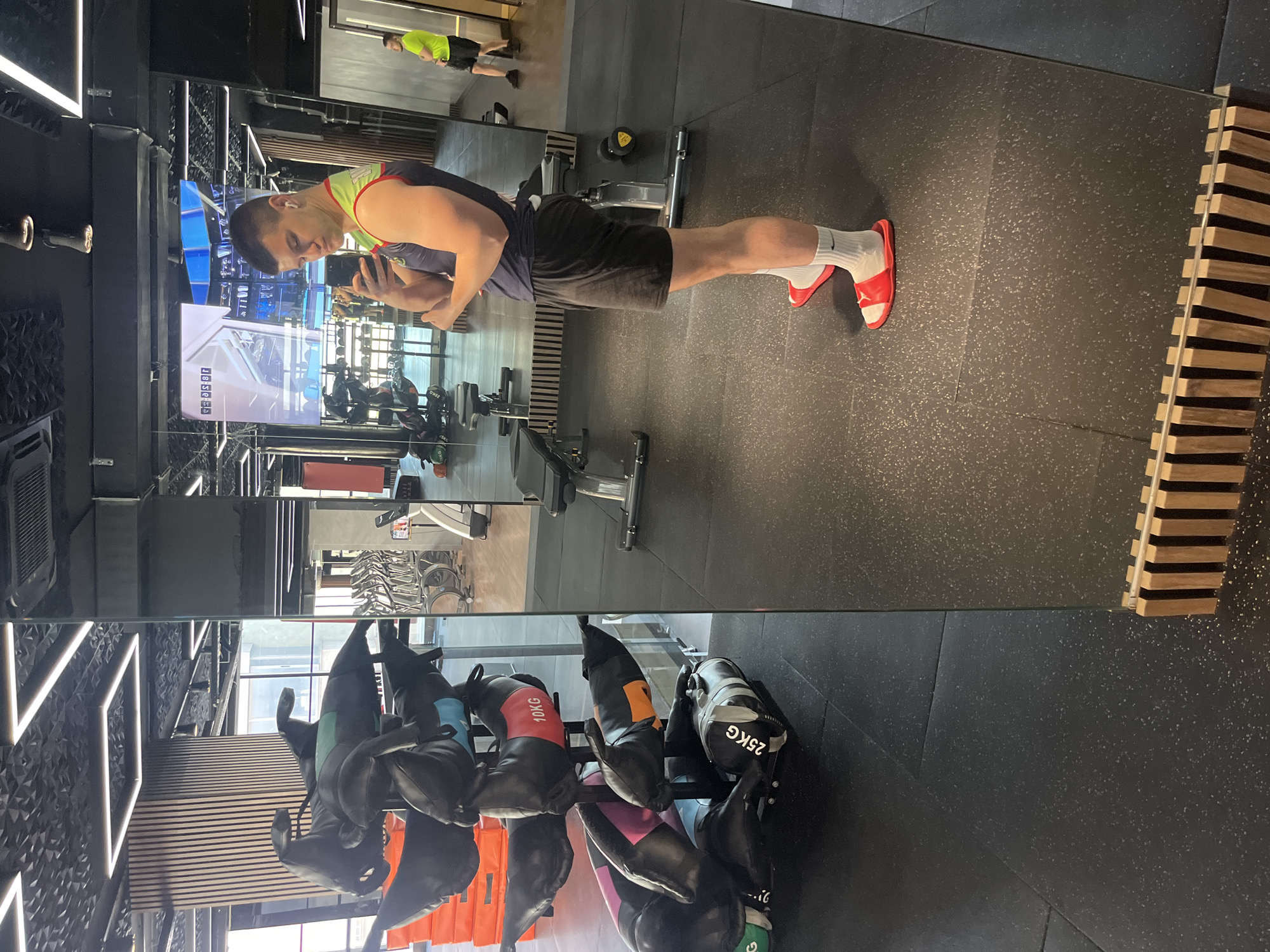 A person is taking a photo of themself in a gym mirror. The individual wears a tank top, shorts, white socks, and red sandals. The gym has exercise equipment, punching bags, and a wooden slat accent on the wall. The location is Sofia, Bulgaria.