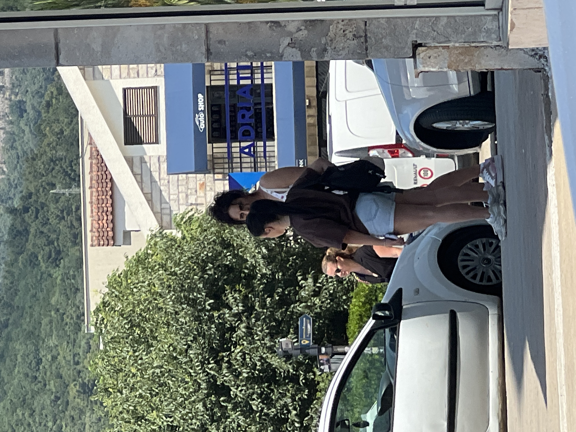 In Budva, Montenegro, on a sunny day, three individuals are present near a street. A woman with blonde hair and sunglasses is standing to the left, wearing a dark top. To her right, a person with dark, curly hair is looking towards the viewer, wearing a white top. Next to them, a young woman with dark hair pulled back is wearing a brown t-shirt and denim shorts, holding a black backpack. They are positioned near a white car, with another white vehicle visible further down the street. Behind them, a building features a blue awning with the text "Auto Shop" and "ADRIA" in large blue letters. A traffic signpost with directional arrows is also visible. The background includes lush green trees and hills, characteristic of the Montenegrin landscape.
